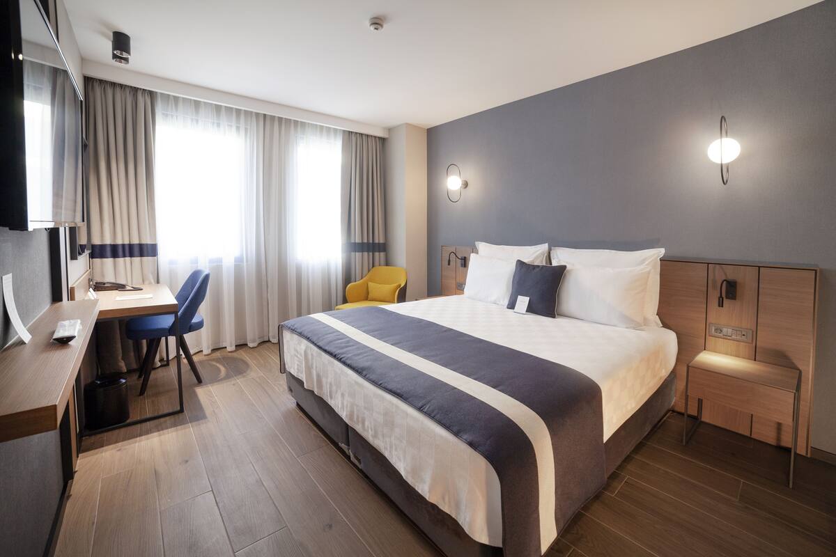 A hotel room with a large bed, a desk with a chair, a small side table, and a TV.