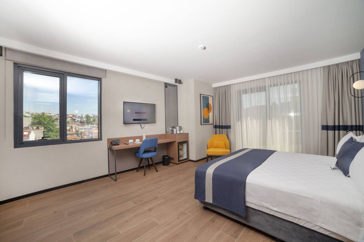 A hotel room with a bed, a desk, a chair, a television, and a window with a city view.