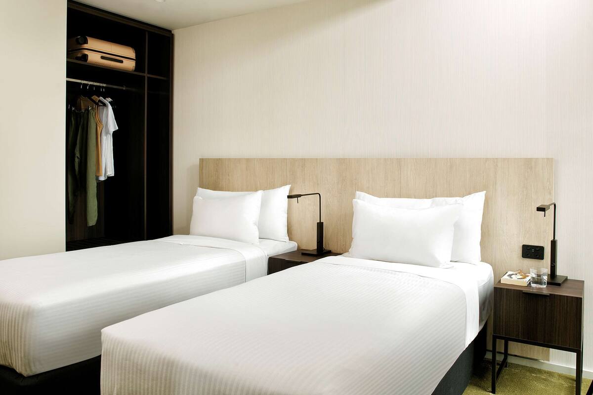 Une chambre d’hôtel avec deux lits simples, une armoire, une table de chevet avec une lampe et une petite table de nuit.