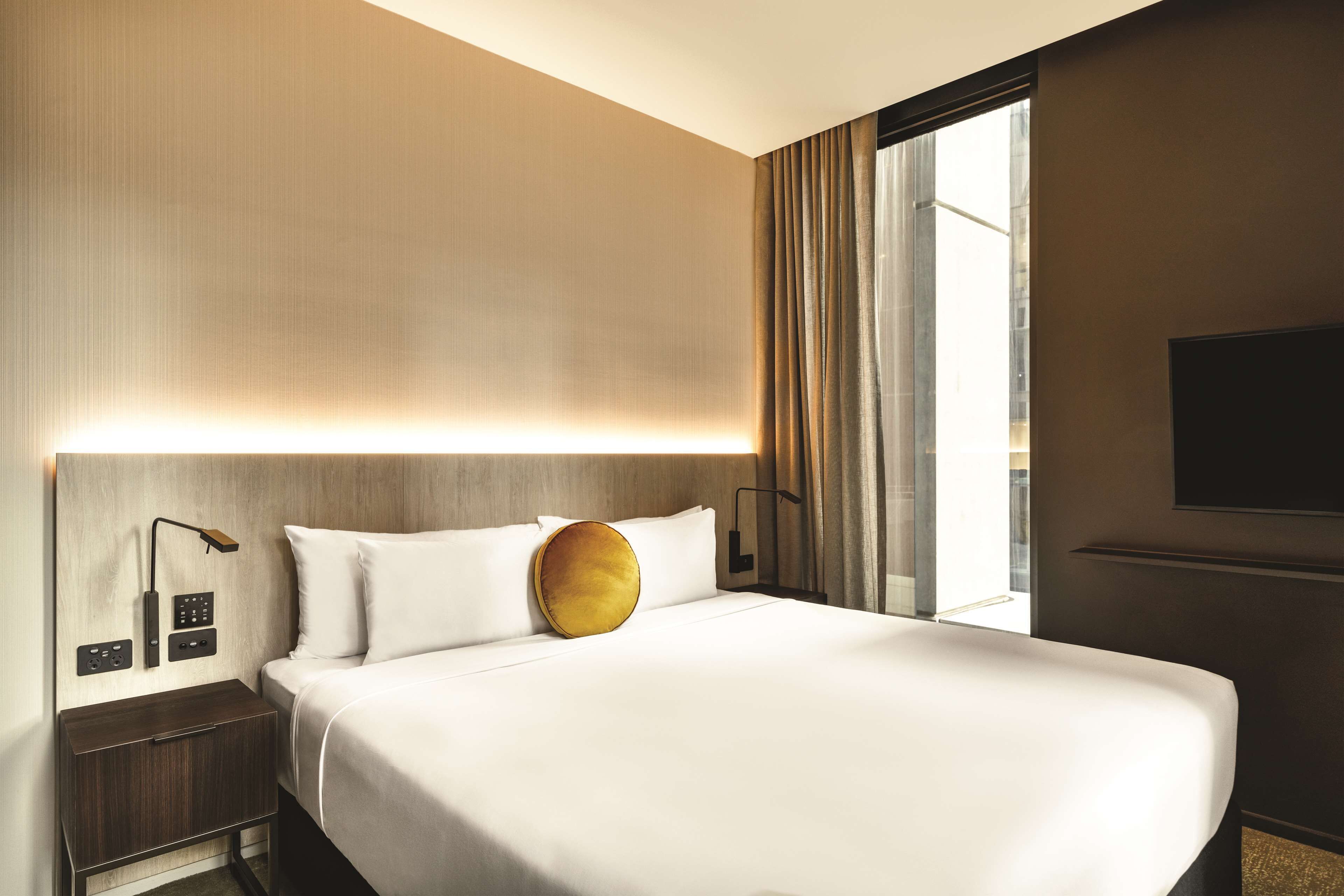 A hotel room with a large bed, a nightstand with a lamp, a flat-screen TV mounted on the wall, and a window with curtains.