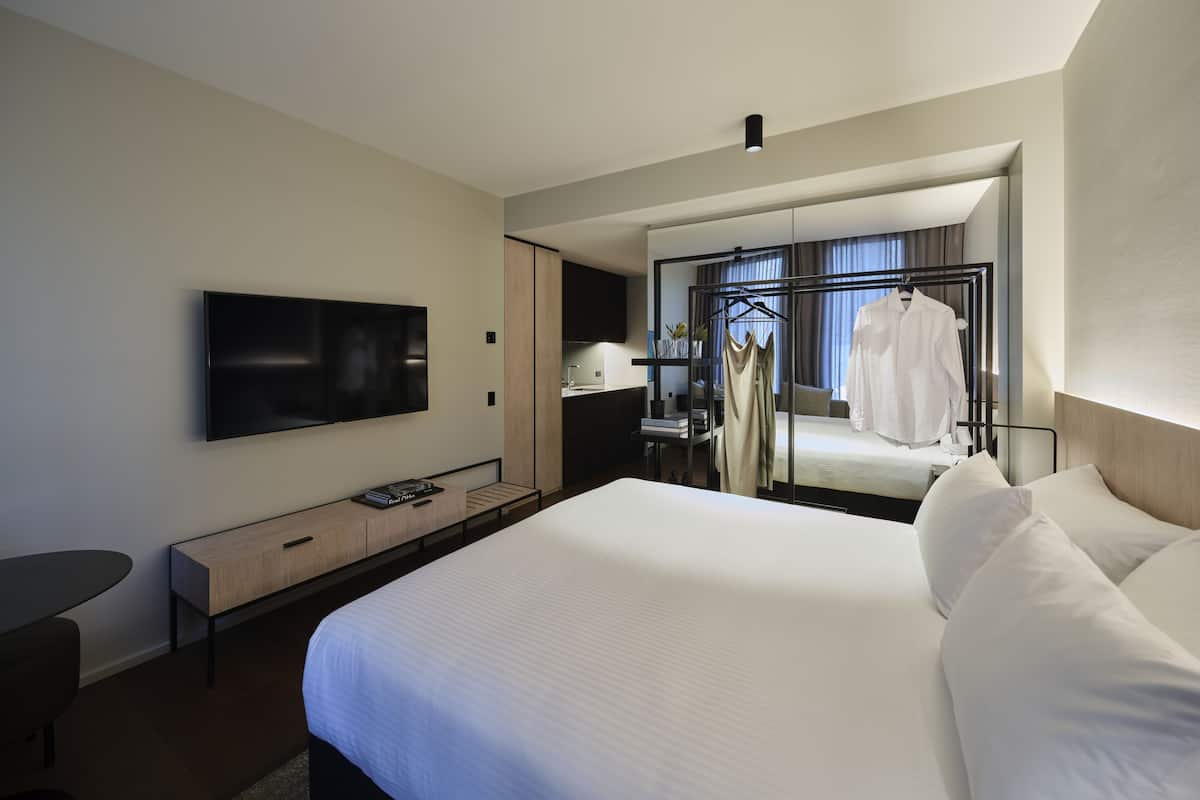 Une chambre d’hôtel moderne avec un grand lit, un téléviseur fixé au mur, un bureau avec une chaise et un miroir qui reflète la pièce.
