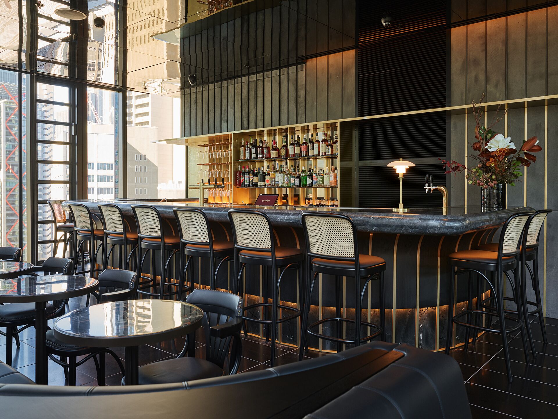 Un espace bar moderne doté d’un haut plafond, de grandes fenêtres et de différents types de sièges, notamment des tables rondes et des tabourets de bar.