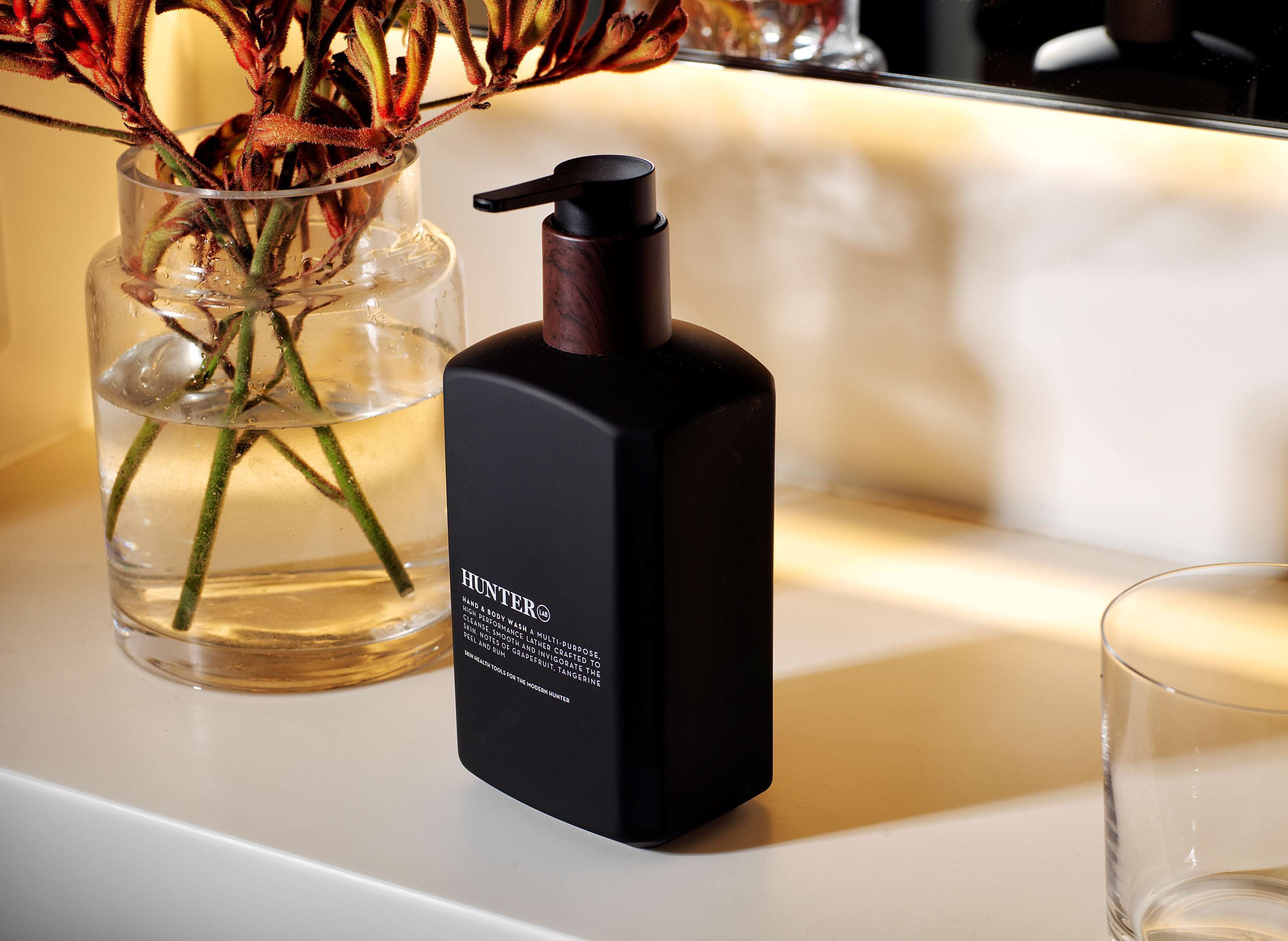 A black hand sanitizer bottle labeled "Hunter" on a white surface next to a clear glass vase with dried flowers.