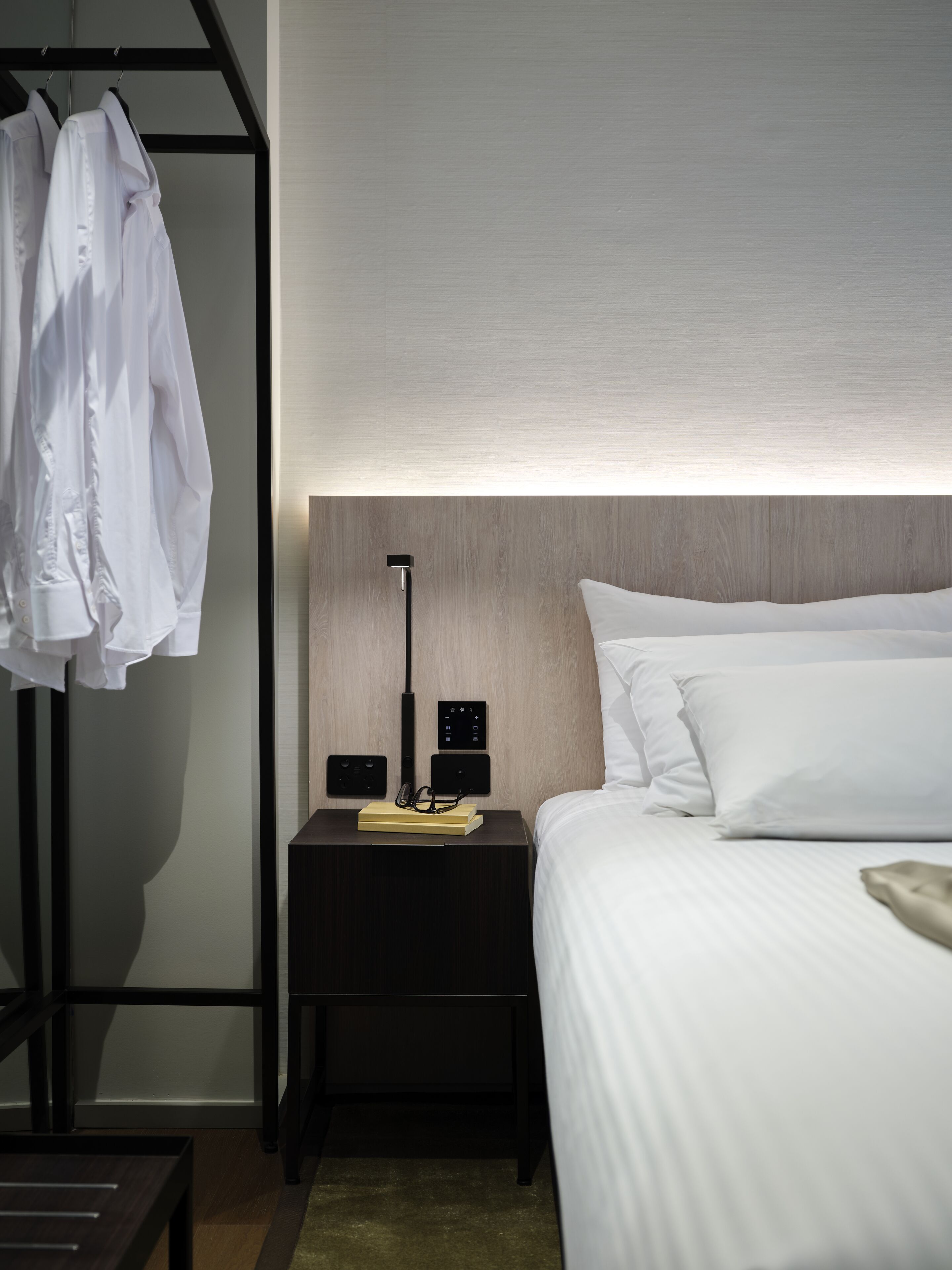 Ett hotellrum med en snyggt bäddad säng, ett sängbord med en lampa och en bok samt en klädställning.
