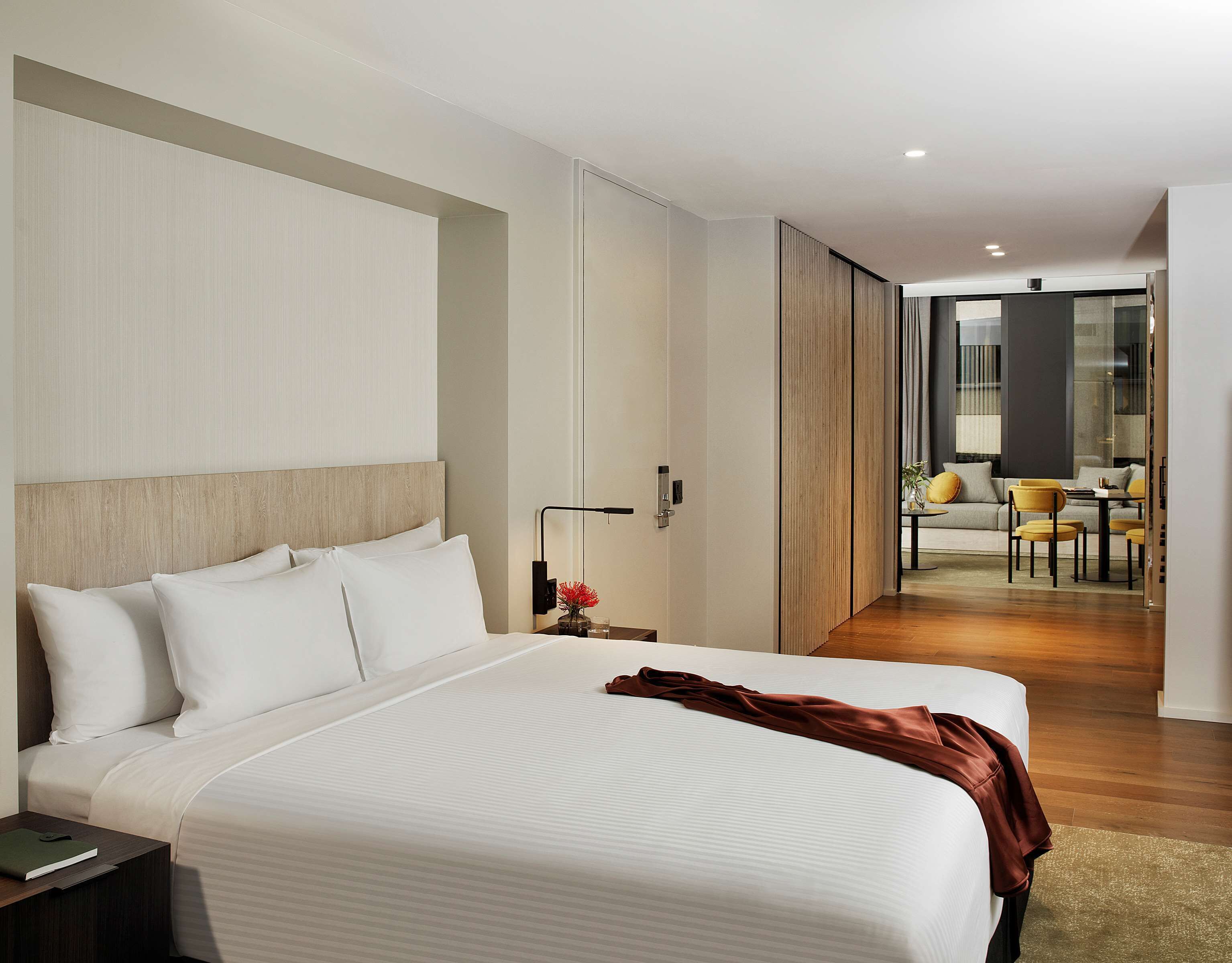 A modern hotel room with a large bed, a bedside table, a nightstand, and a view into the living area.