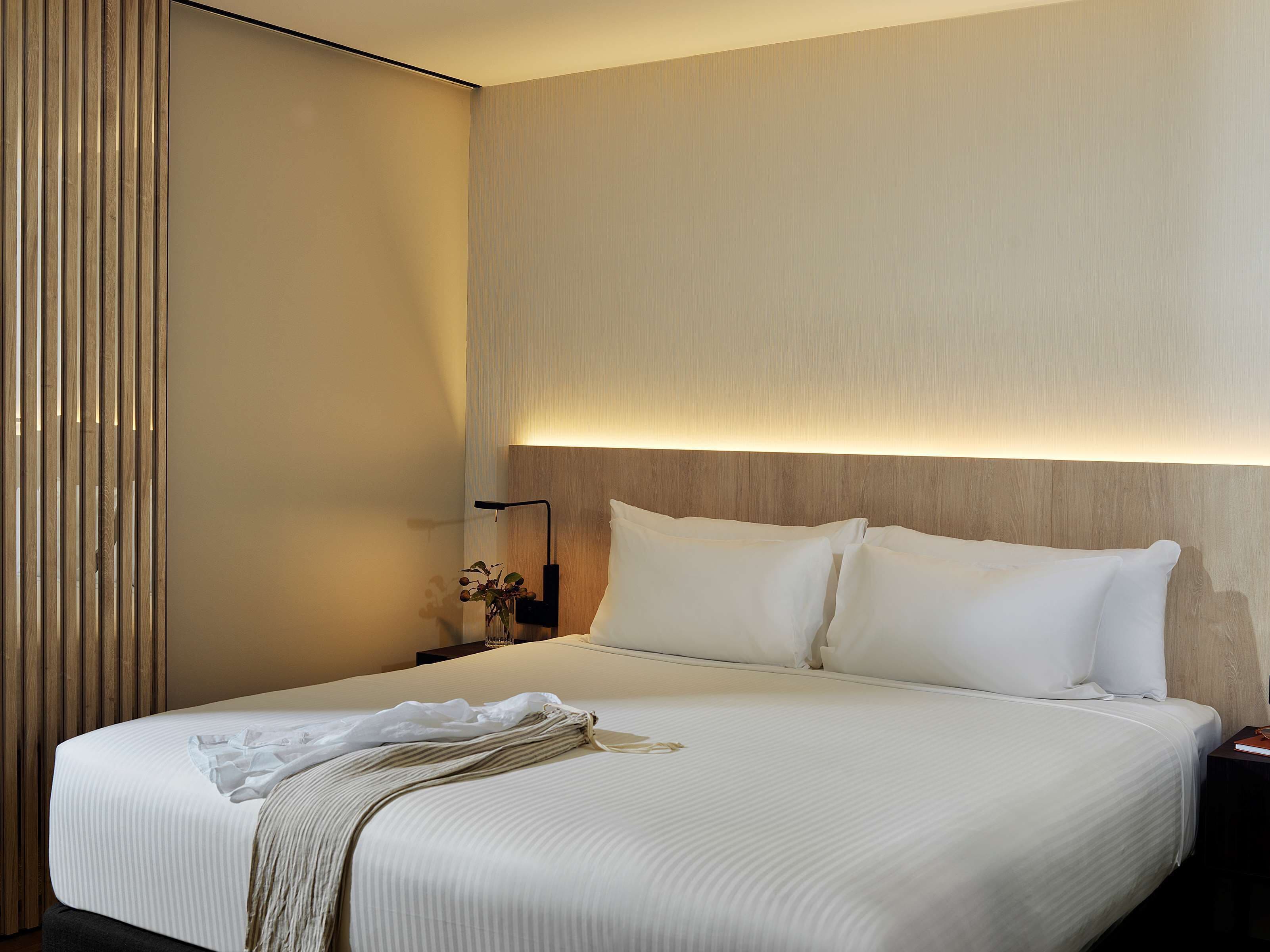 A neatly made bed with white linens, a bedside table with a lamp, and a wooden panel on the left.