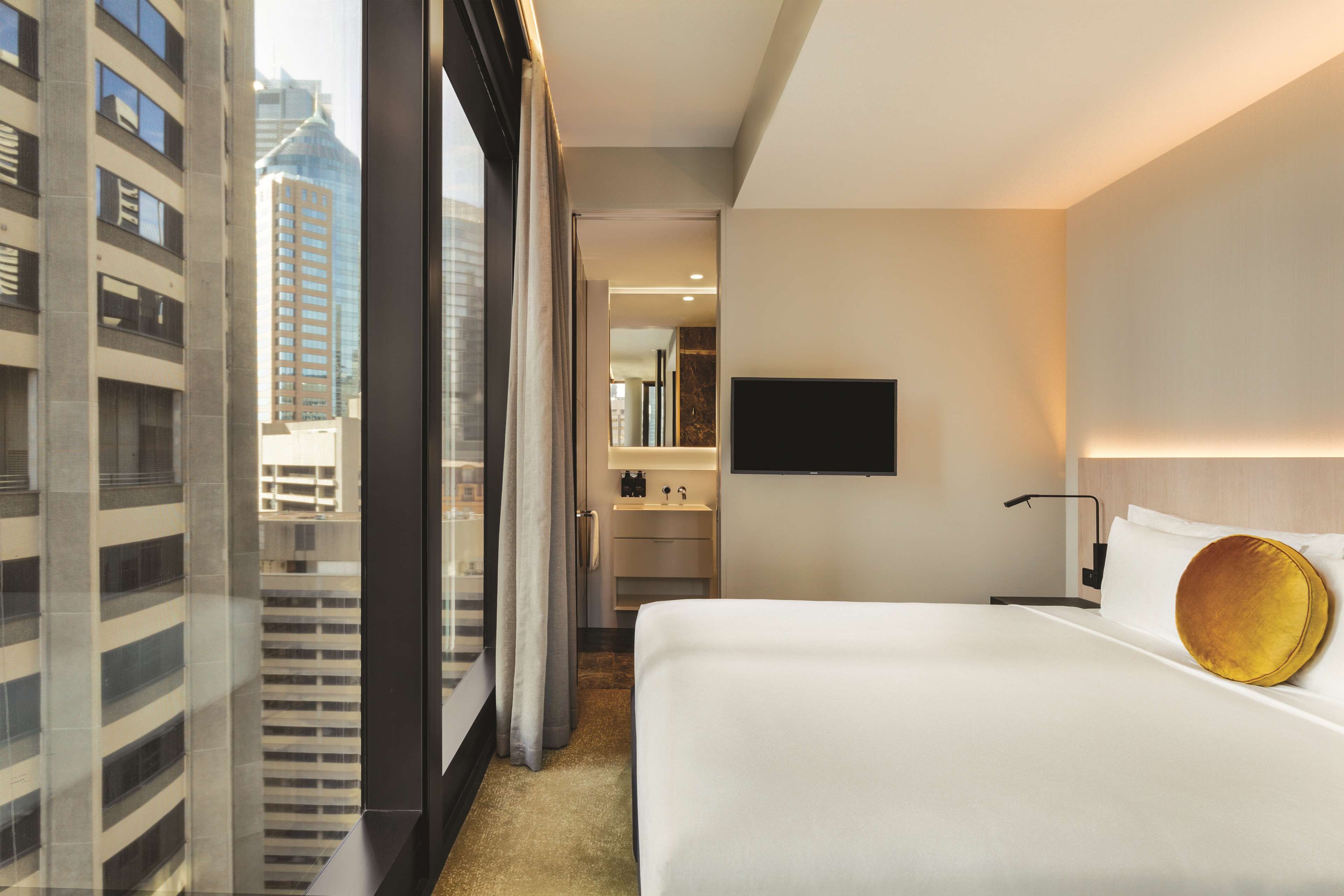 A hotel room with a large bed, a TV mounted on the wall, and a view of tall buildings through the window.