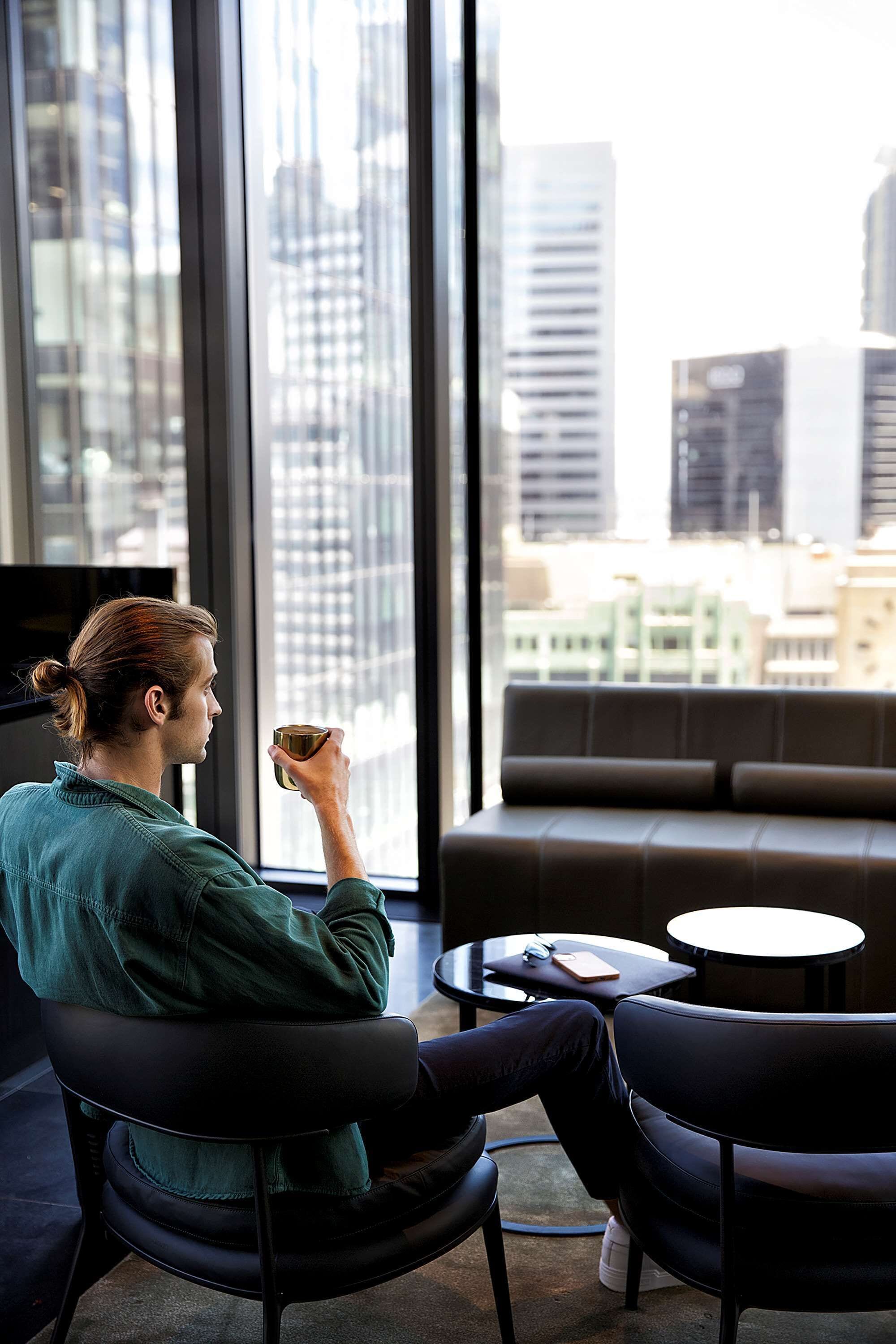 A person sitting on a chair by a window with a city view, holding a cup.