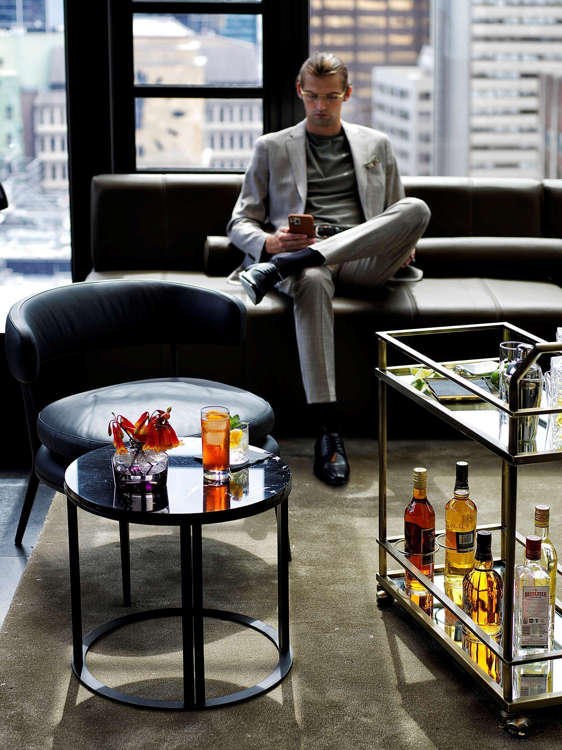 A man sitting on a leather couch in a modern room with a city view, a round table with a drink and flowers, and a bar cart with bottles.