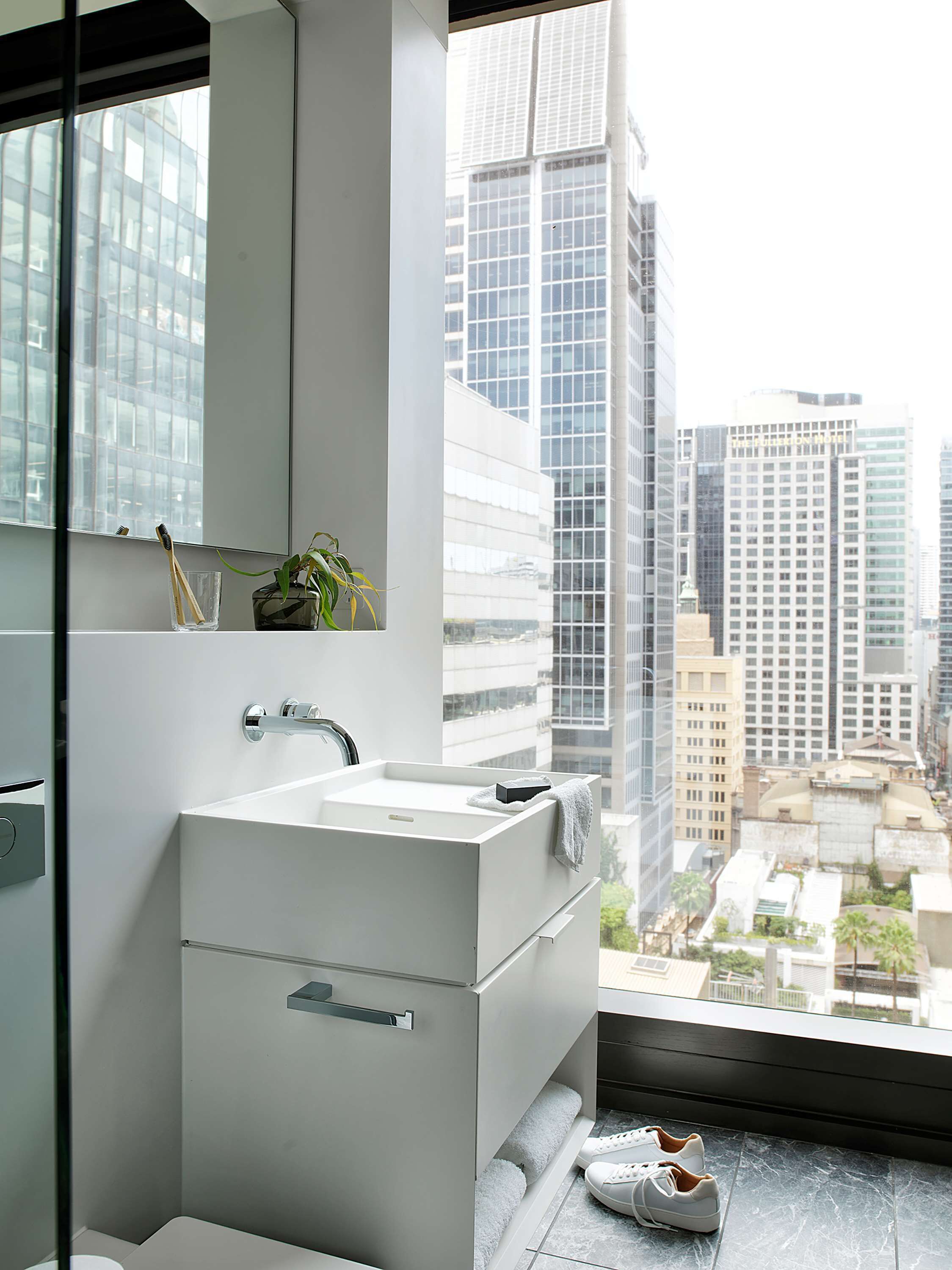 A modern bathroom with a white sink, a mirror, and a view of a cityscape through a large window.