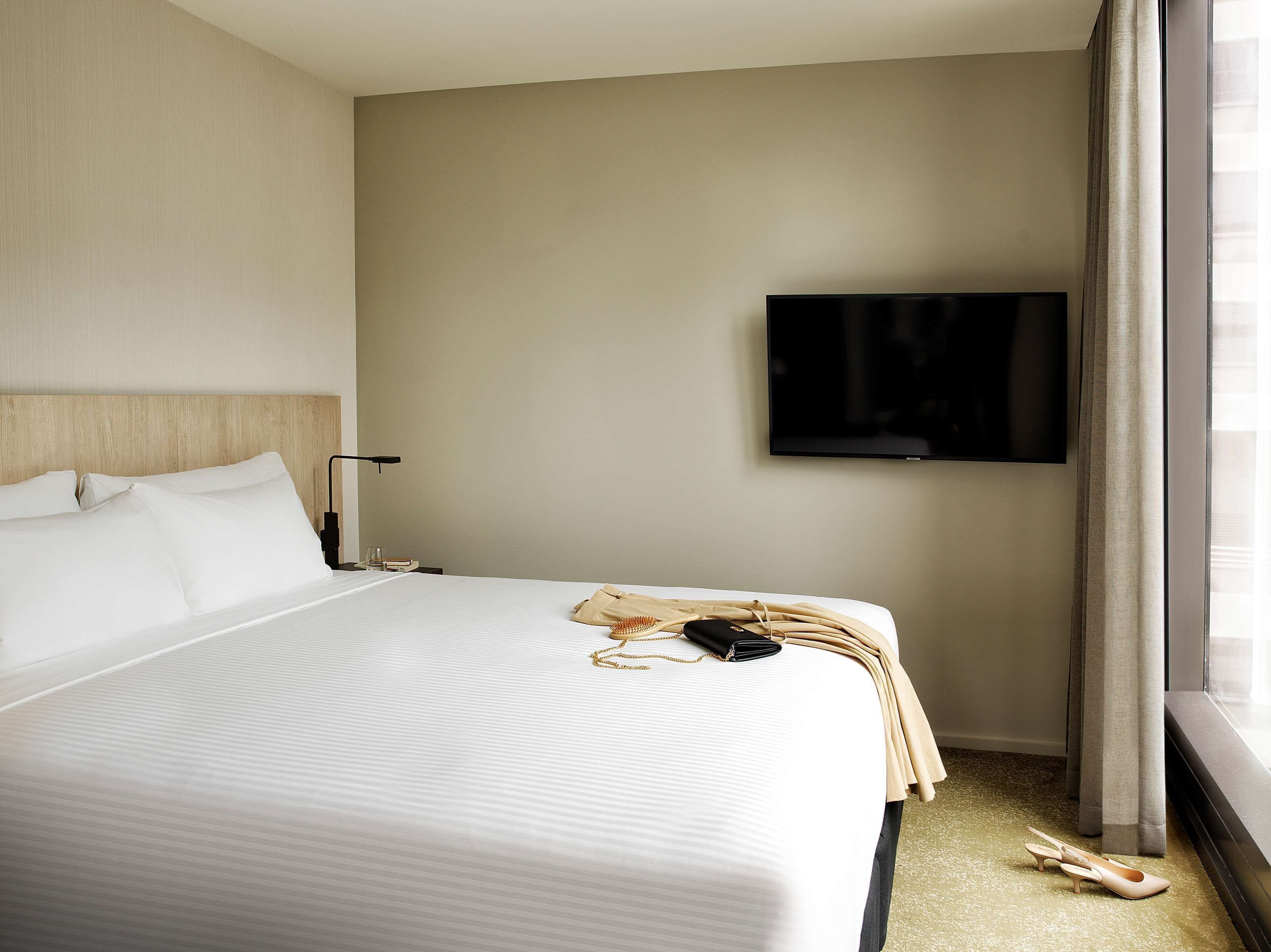 A hotel room with a large bed, a flat-screen TV mounted on the wall, and a window with curtains.