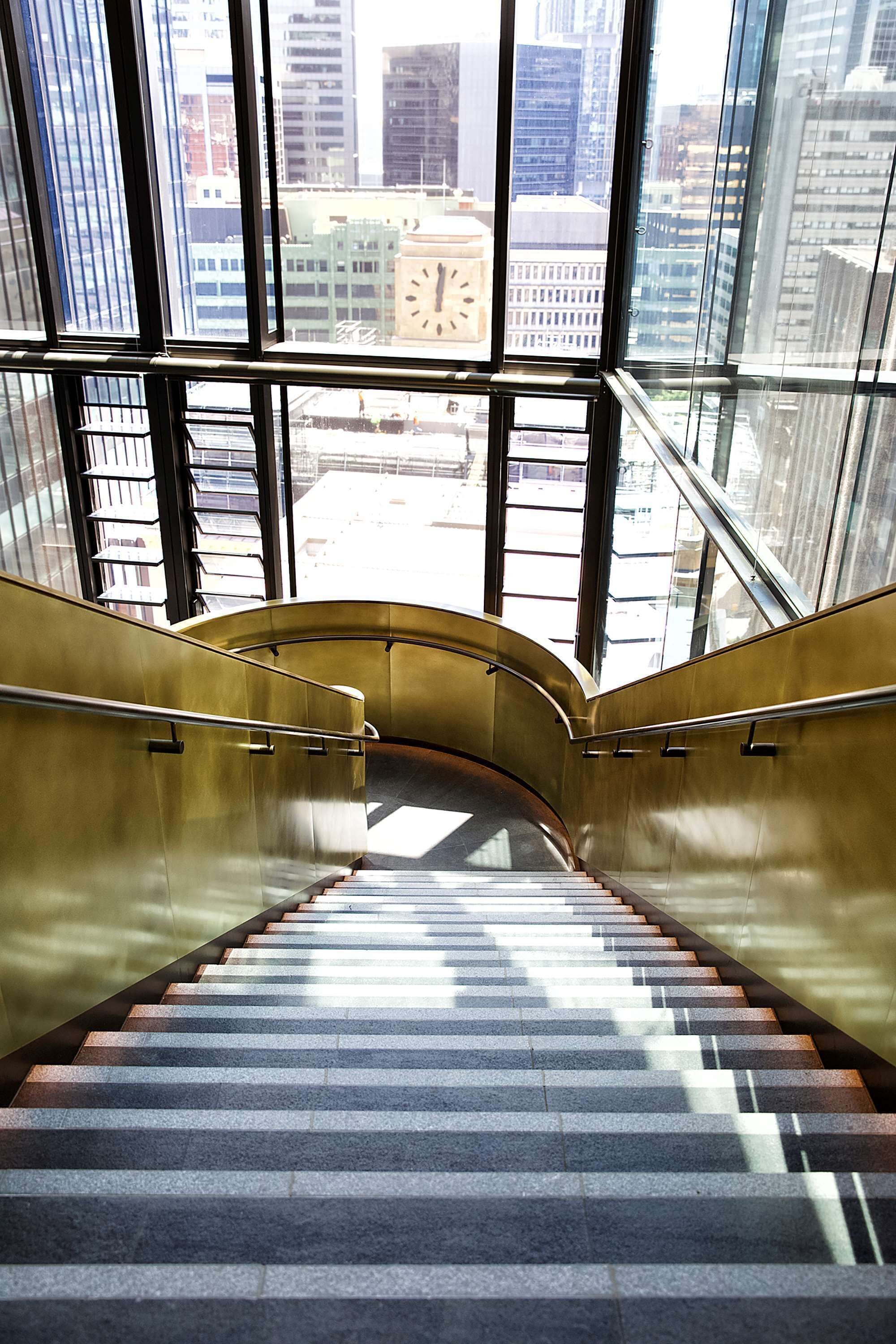 Una escalera con barandas de cristal y vistas a un paisaje urbano a través de amplios ventanales.