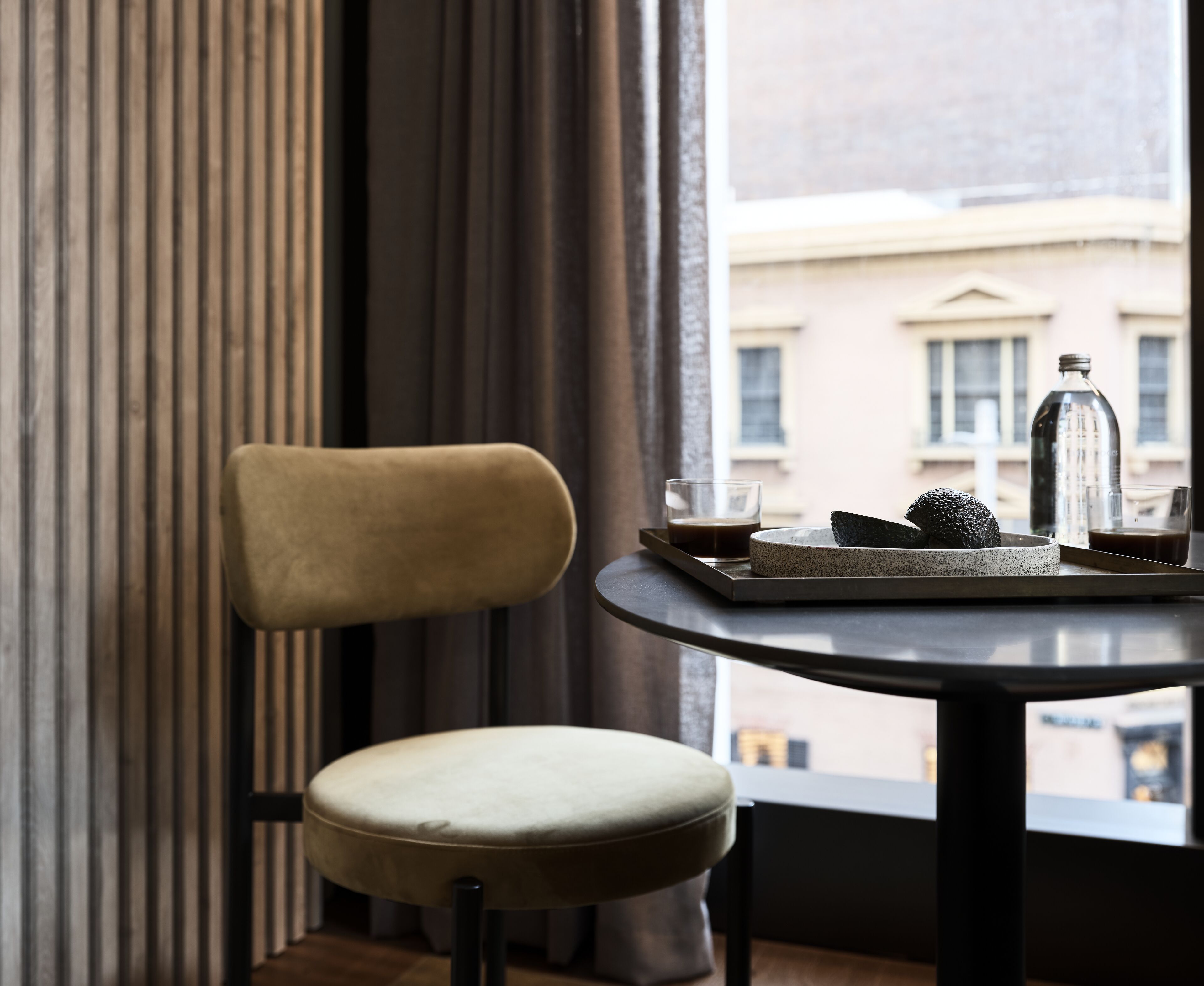 A chair and table with a glass of coffee and a bottle, near a window with a view of buildings.