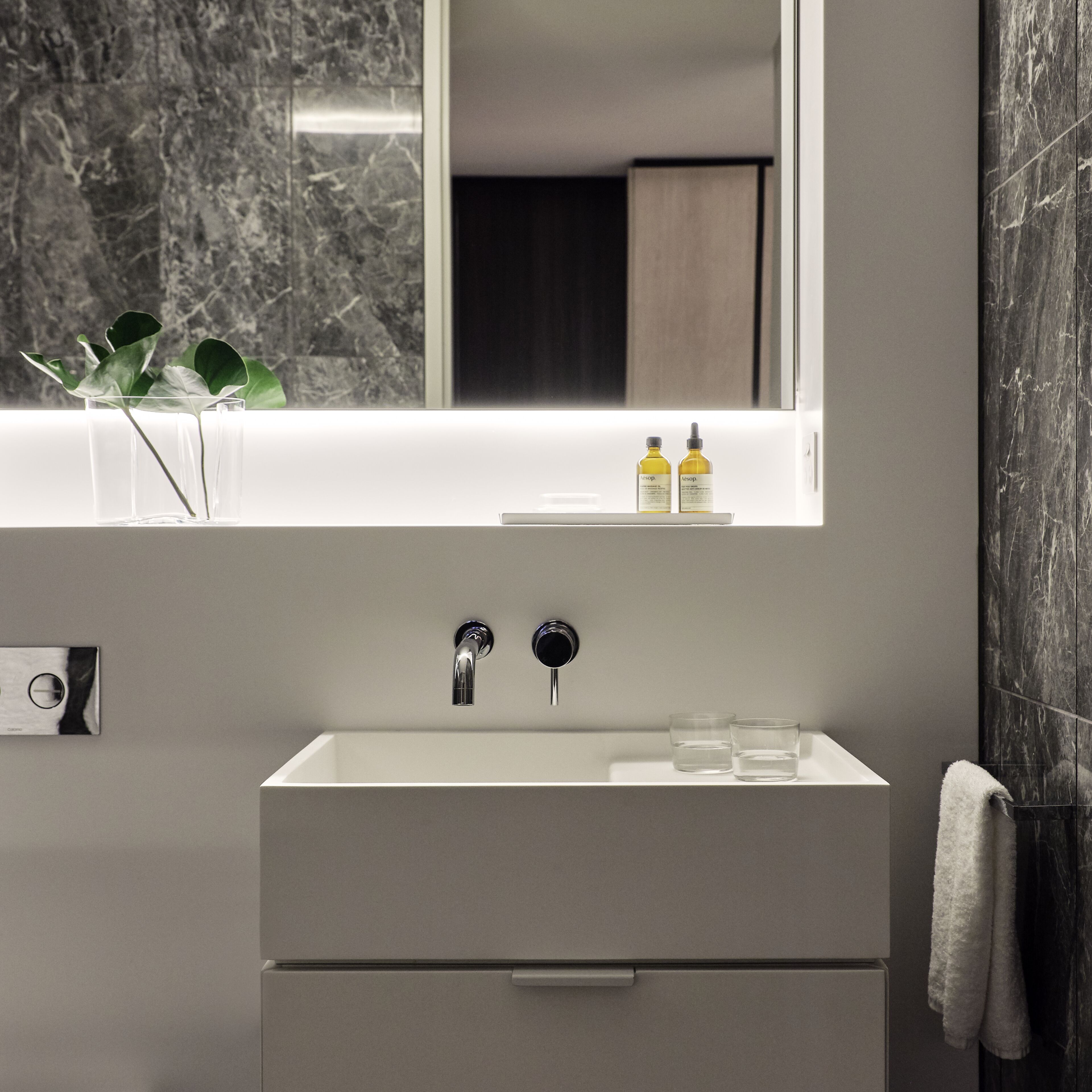 A modern bathroom with a white sink, two faucets, a mirror above the sink, and a window with a view of a plant.