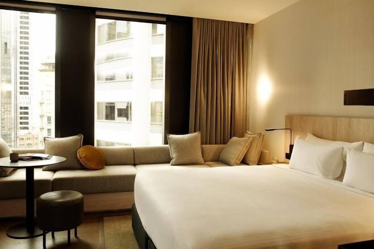 Une chambre d’hôtel avec un grand lit, un canapé, une petite table et une vue sur la ville grâce à de grandes fenêtres.