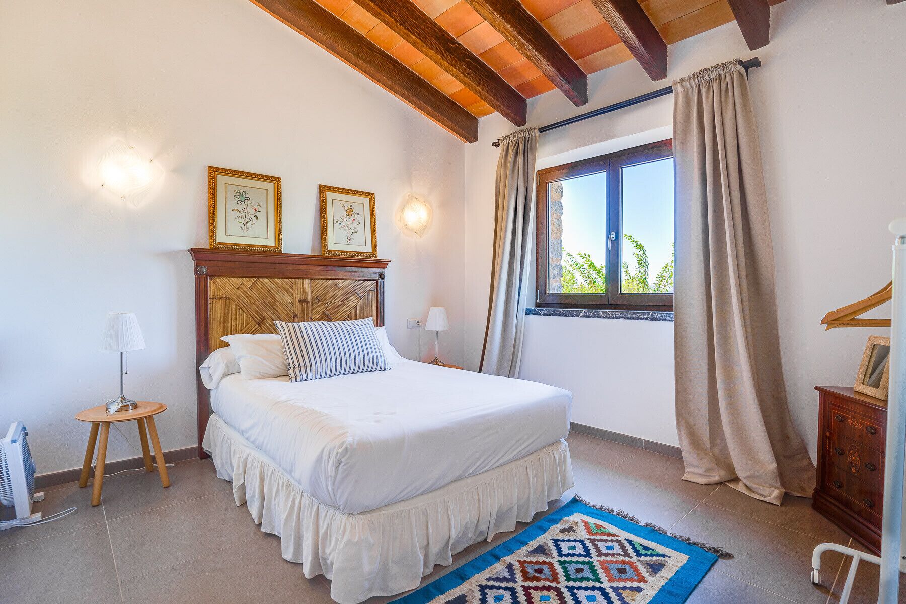 Une chambre à coucher avec un lit, une fenêtre avec des rideaux, une table de chevet, une lampe et un luminaire fixé au mur.