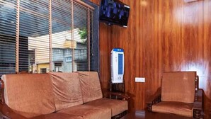 A wooden-furnished living area with a sofa and a table, a television mounted on the wall, and a window with blinds. - Swarnam Residency (Chennai)