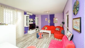 Una sala de estar con un sofá rojo, una mesa de centro blanca y un televisor. Hay una bandera en el suelo y un reloj en la pared.