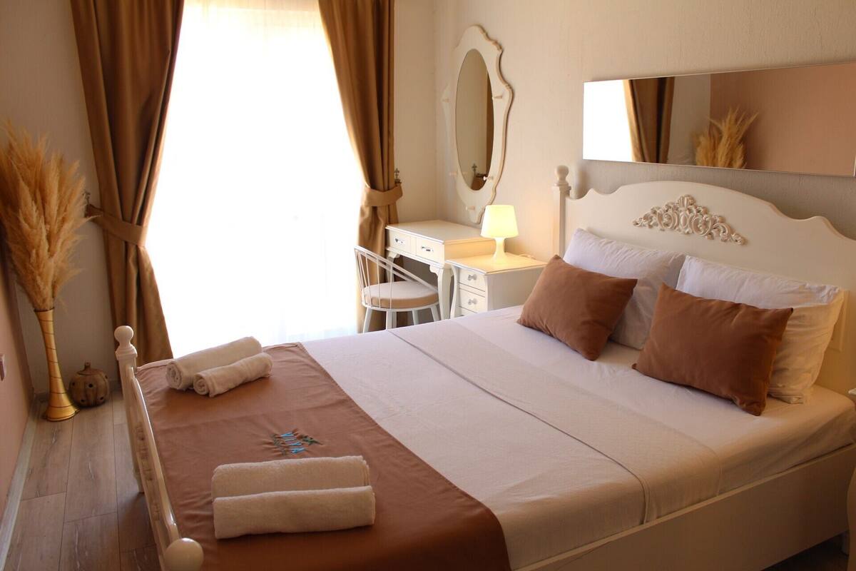 Una cama bien hecha con almohadas y toallas, una mesita de noche con una lámpara, un espejo y una silla en una habitación bien iluminada.
