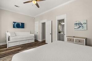 Un dormitorio con una cama blanca, un ventilador de techo, un cuadro y un baño con un espejo y un perchero para toallas.
