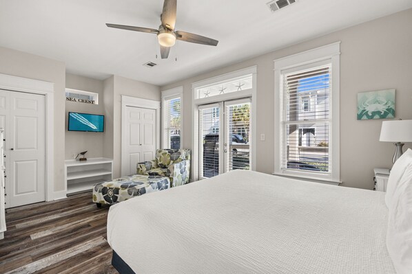 A bedroom with a large bed, a ceiling fan, a TV, and a view of the outside.