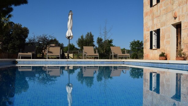 A clear blue pool with lounge chairs and an umbrella, surrounded by stone buildings and trees. - Punta de Vistalegre is a Villa with a Private Pool in a Very Quiet Rural Setting (Porto Cristo)