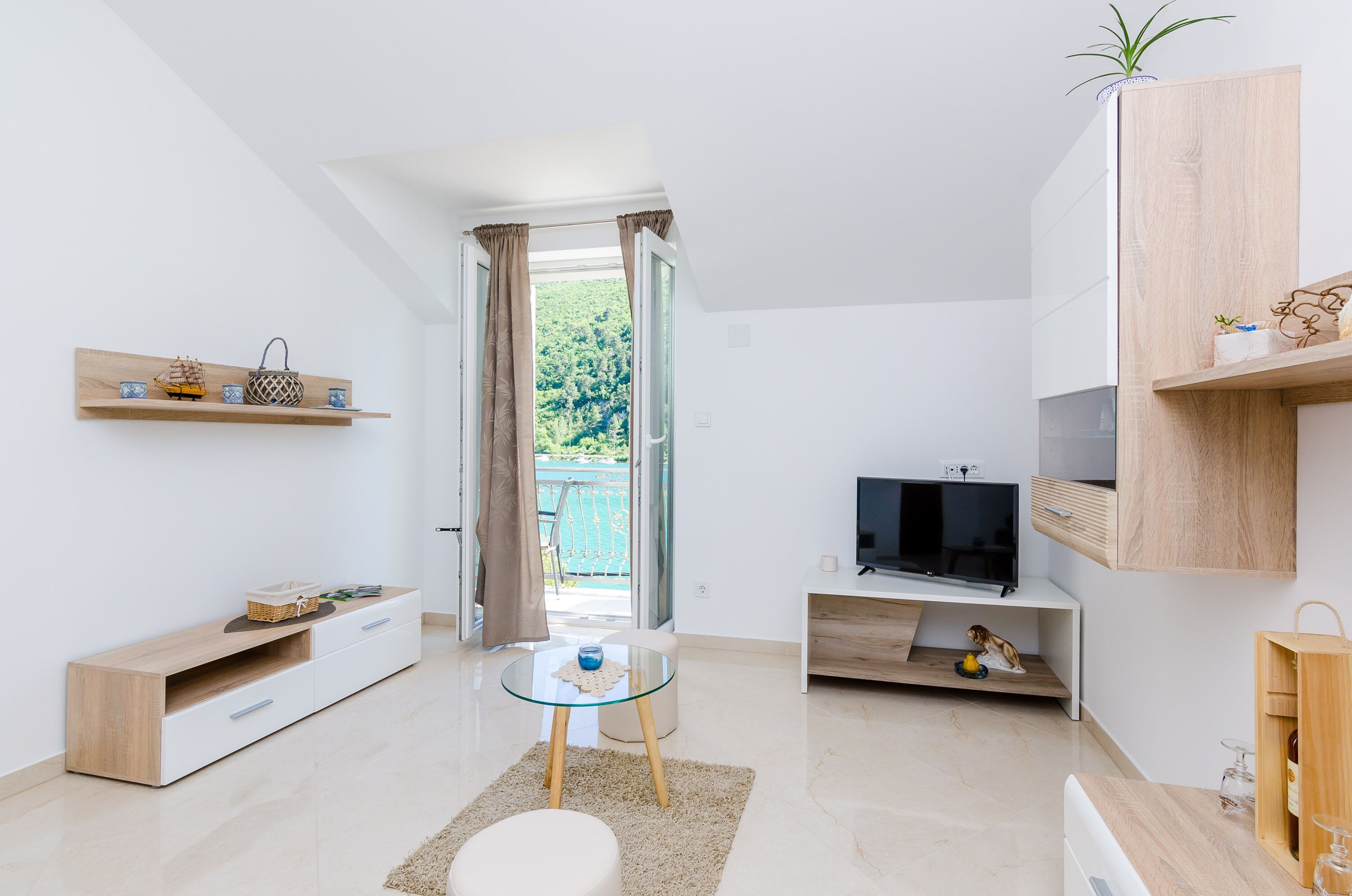 A modern living room with a TV, wooden furniture, a glass table, and a view of a green landscape through an open door.