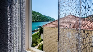 A view from a window overlooking a coastal area with a clear blue sea, a hilly landscape, and a building with a red-tiled roof.
