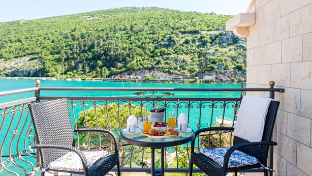 A balcony with two wicker chairs, a small round table with breakfast items, and a view of a turquoise sea and green hills.