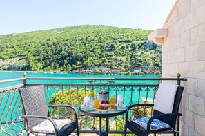 A balcony with two wicker chairs, a small round table with breakfast items, and a view of a turquoise sea and green hills. - Apartments La Luna (Dubrovnik)