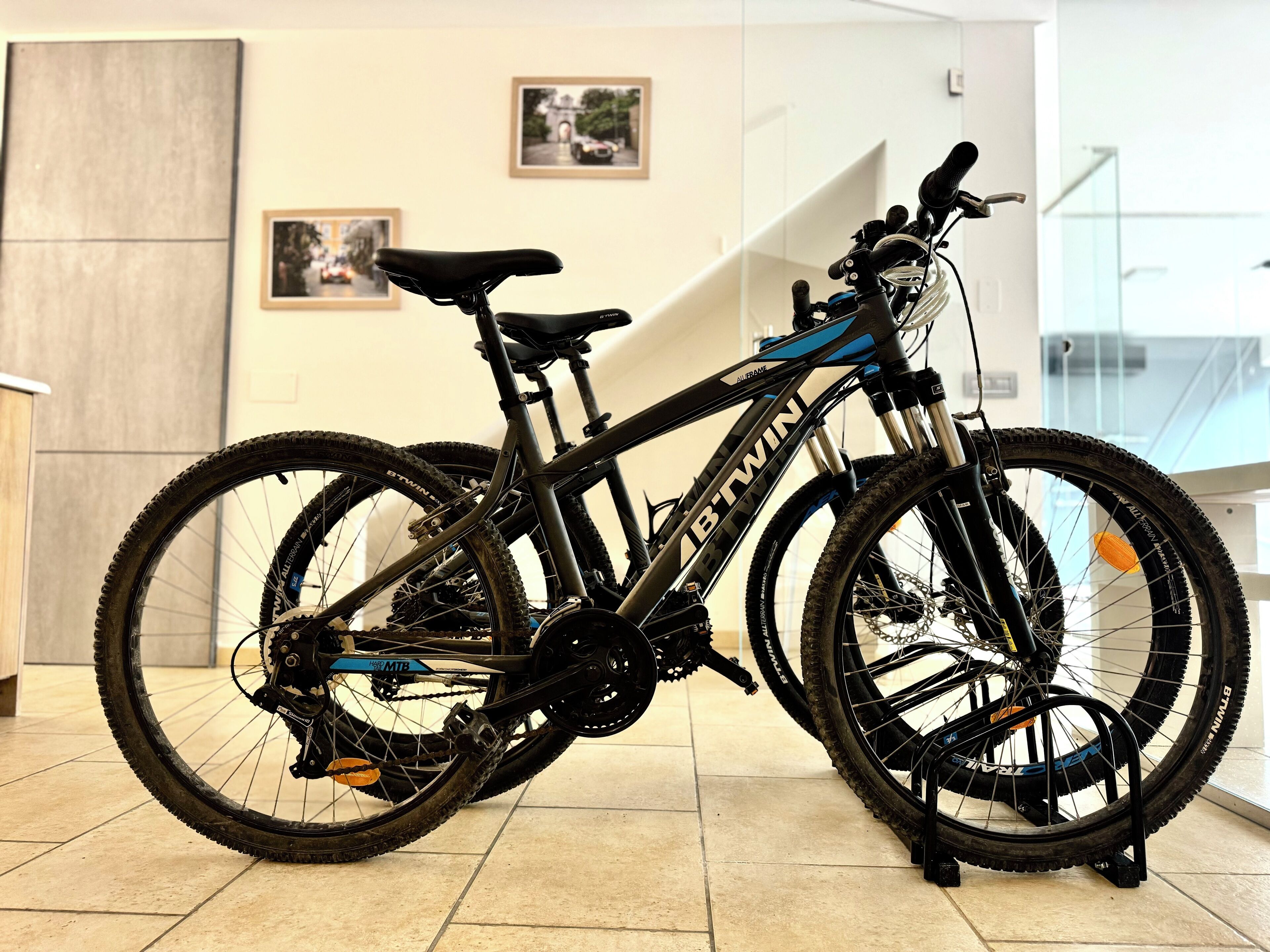 A mountain bike with a black frame and blue accents, equipped with a front suspension fork and a rear derailleur, parked indoors on a tiled floor.