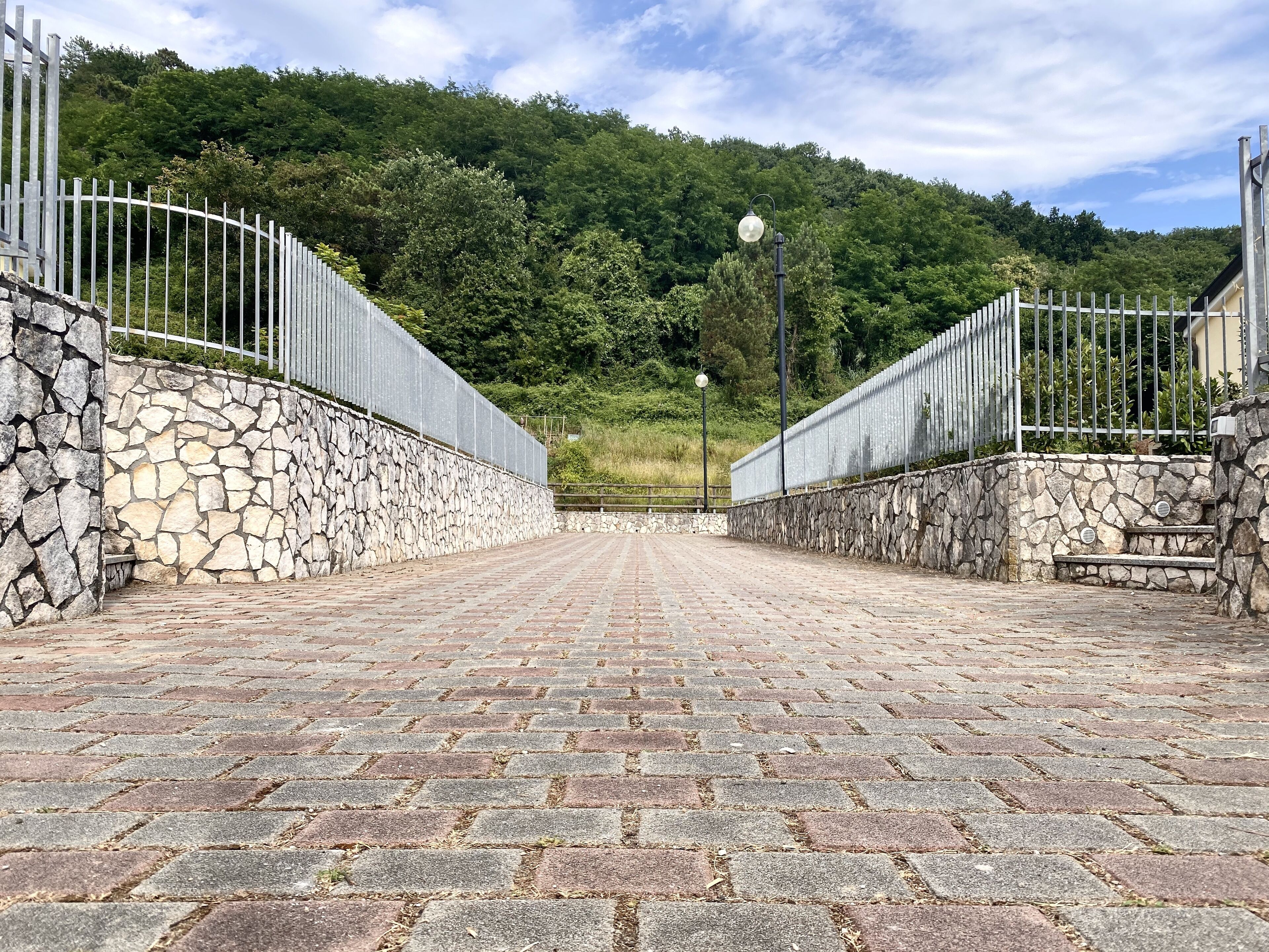 Ein gepflasterter Weg führt zu einer Steinmauer mit Metallzaun an beiden Seiten, umgeben von üppiger Vegetation und einem Hügel im Hintergrund.