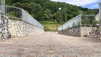 Ein gepflasterter Weg führt zu einer Steinmauer mit Metallzaun an beiden Seiten, umgeben von üppiger Vegetation und einem Hügel im Hintergrund.