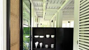 Un pasillo con un estante negro que contiene platos y cuencos blancos, un techo con tuberías a la vista y vistas de follaje exterior.