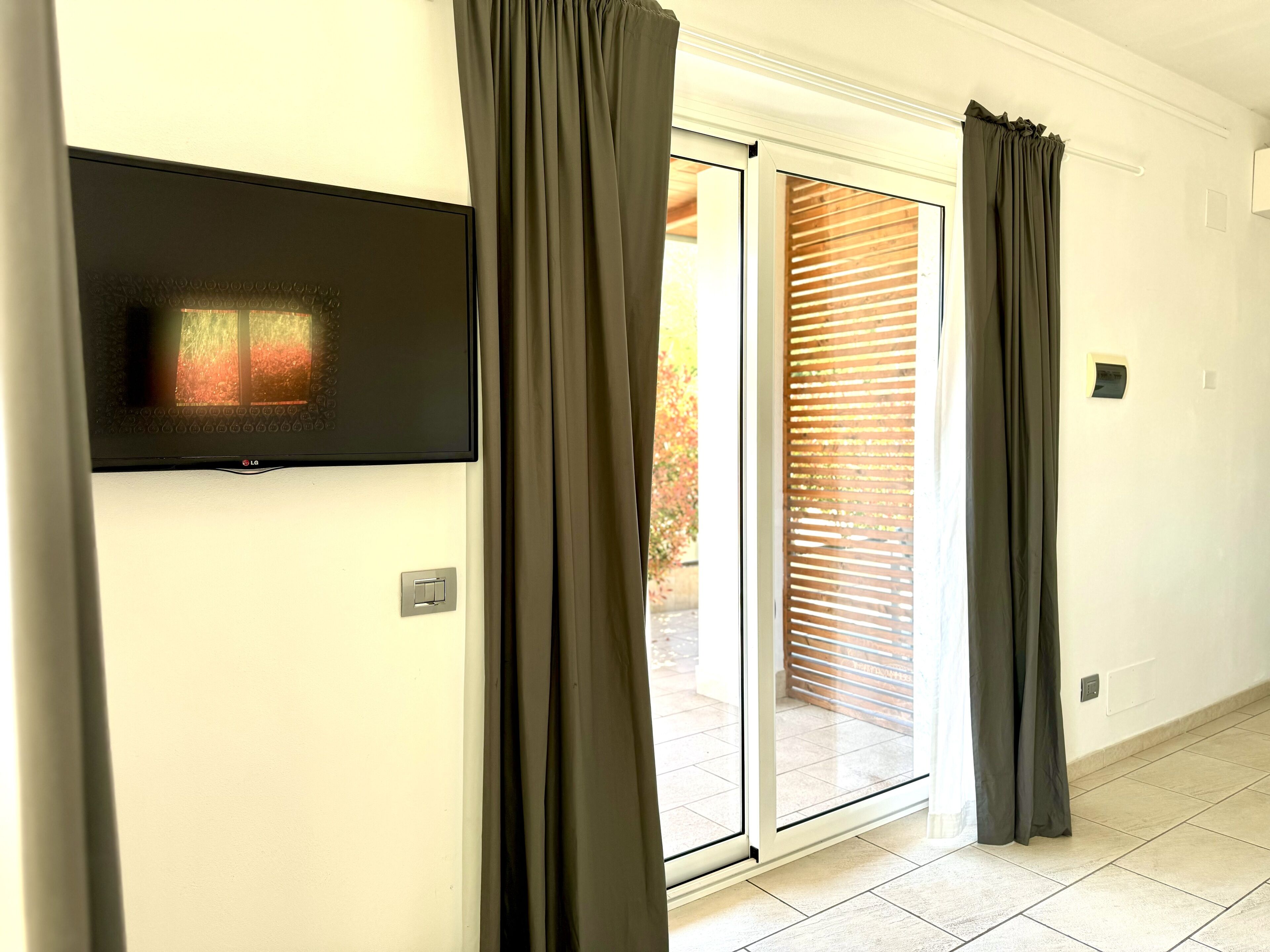A room with a sliding glass door, a flat-screen TV mounted on the wall, and a light switch.