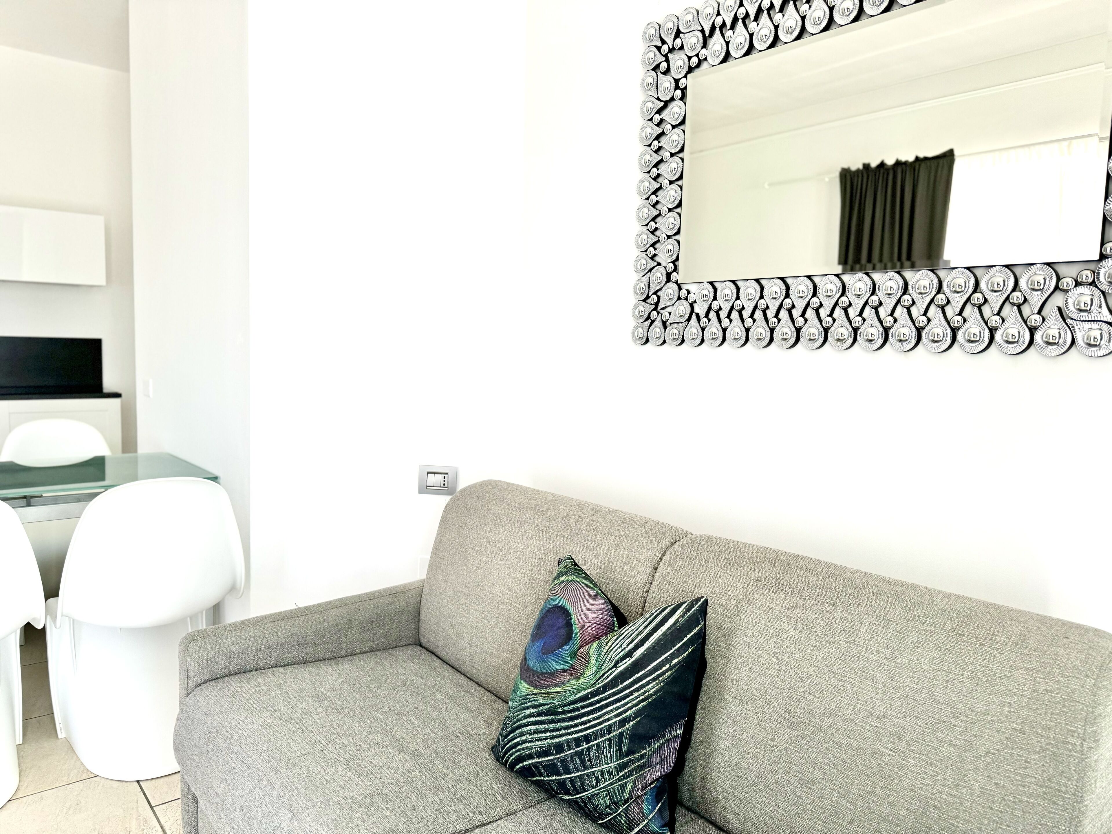 A modern living room with a grey sofa, a colorful pillow, a black-framed mirror, and a dining area with white chairs and a glass table.