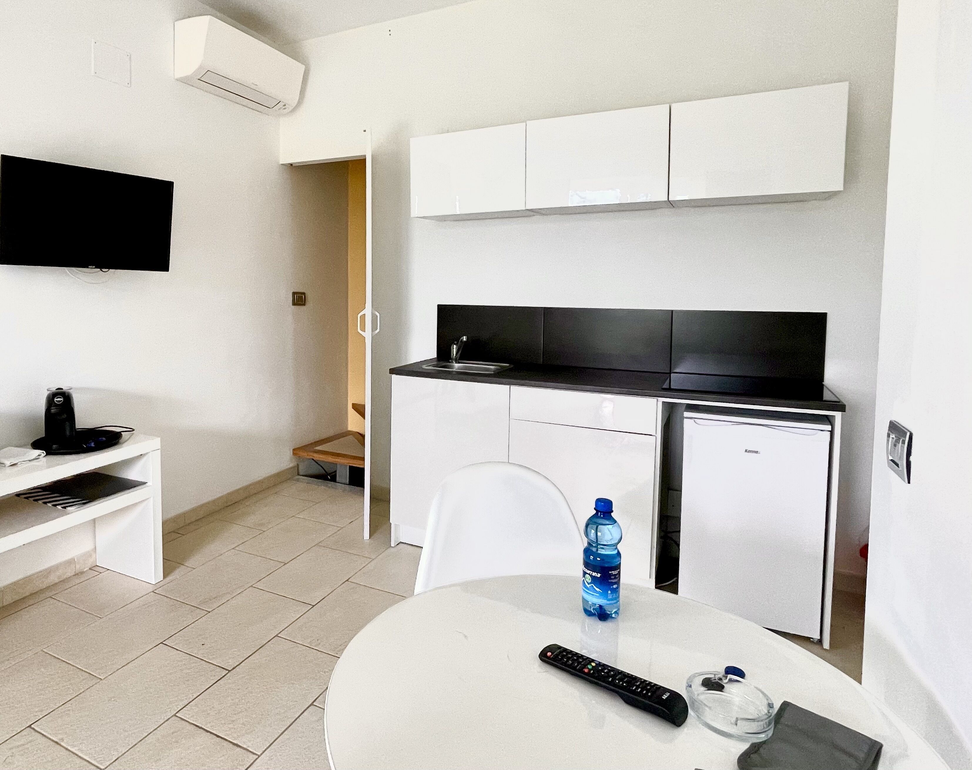 A compact kitchen with white cabinets, a black countertop, and a small refrigerator. A water bottle and a remote control are on the table.
