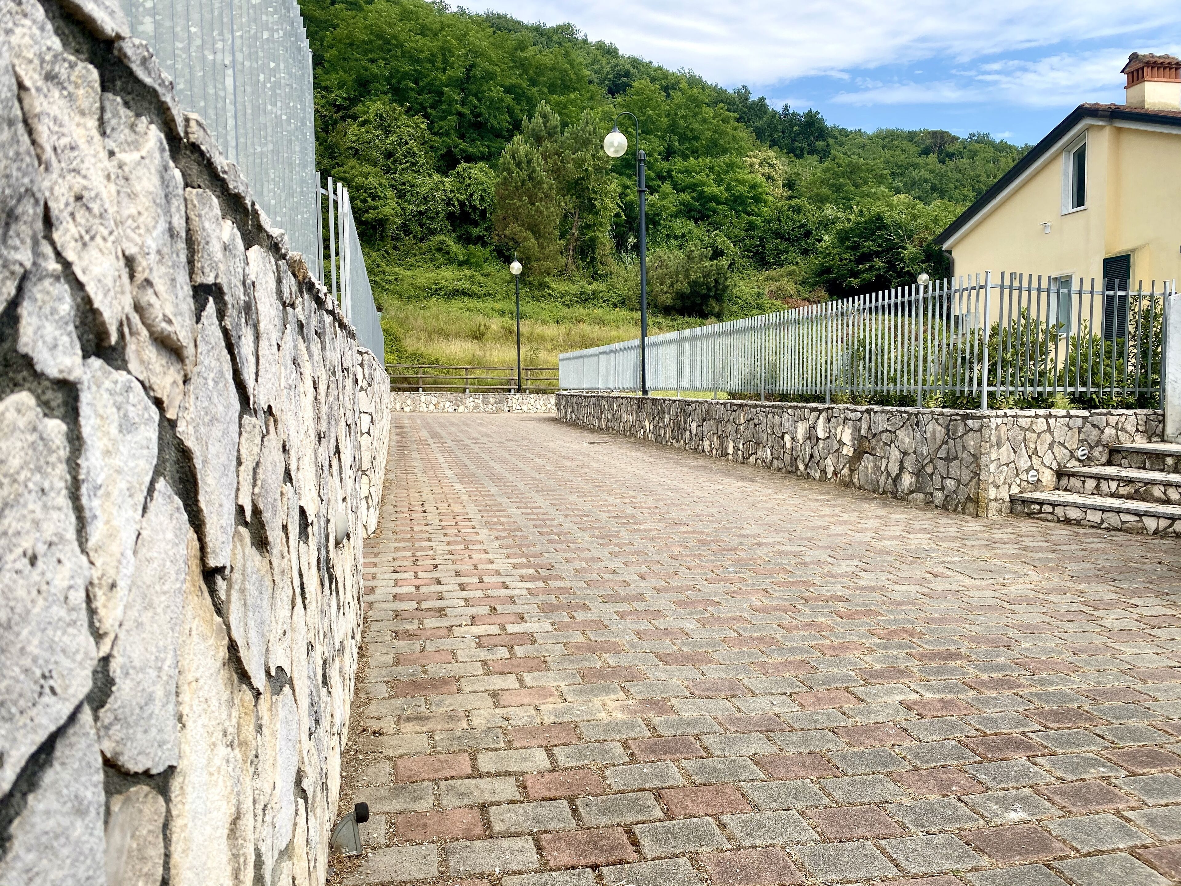Un chemin pavé mène à un mur de pierre et à un bâtiment entouré d’une clôture en métal.