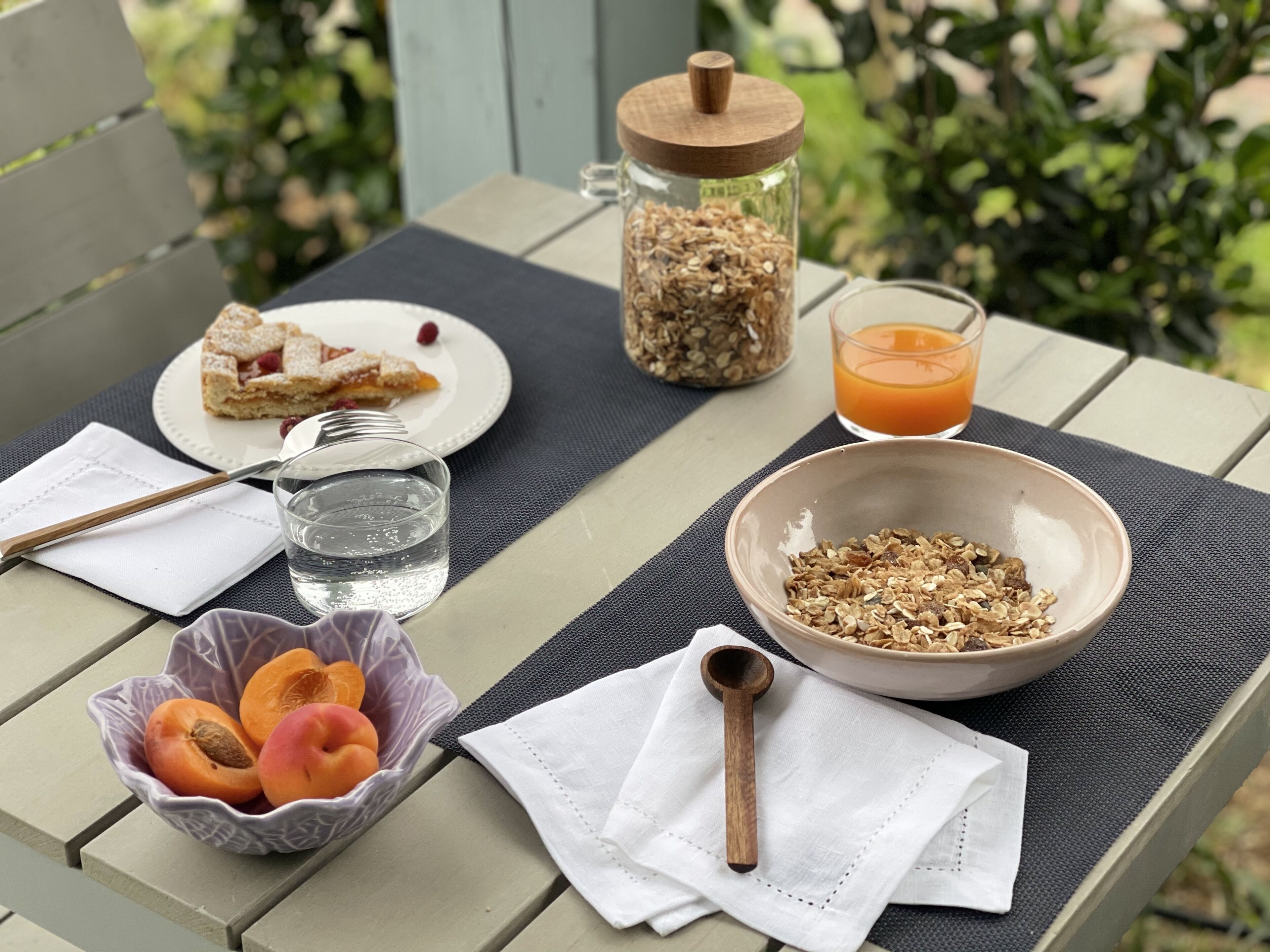 Ein Frühstückstisch im Freien mit einem Glas Orangensaft, einer Schüssel Granola, einem Teller mit Gebäck, einem Einmachglas mit Saaten und einer Obstschale.