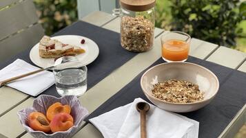 A breakfast setup on an outdoor table with a glass of orange juice, a bowl of granola, a plate with a pastry, a jar of seeds, and a bowl of fruit.