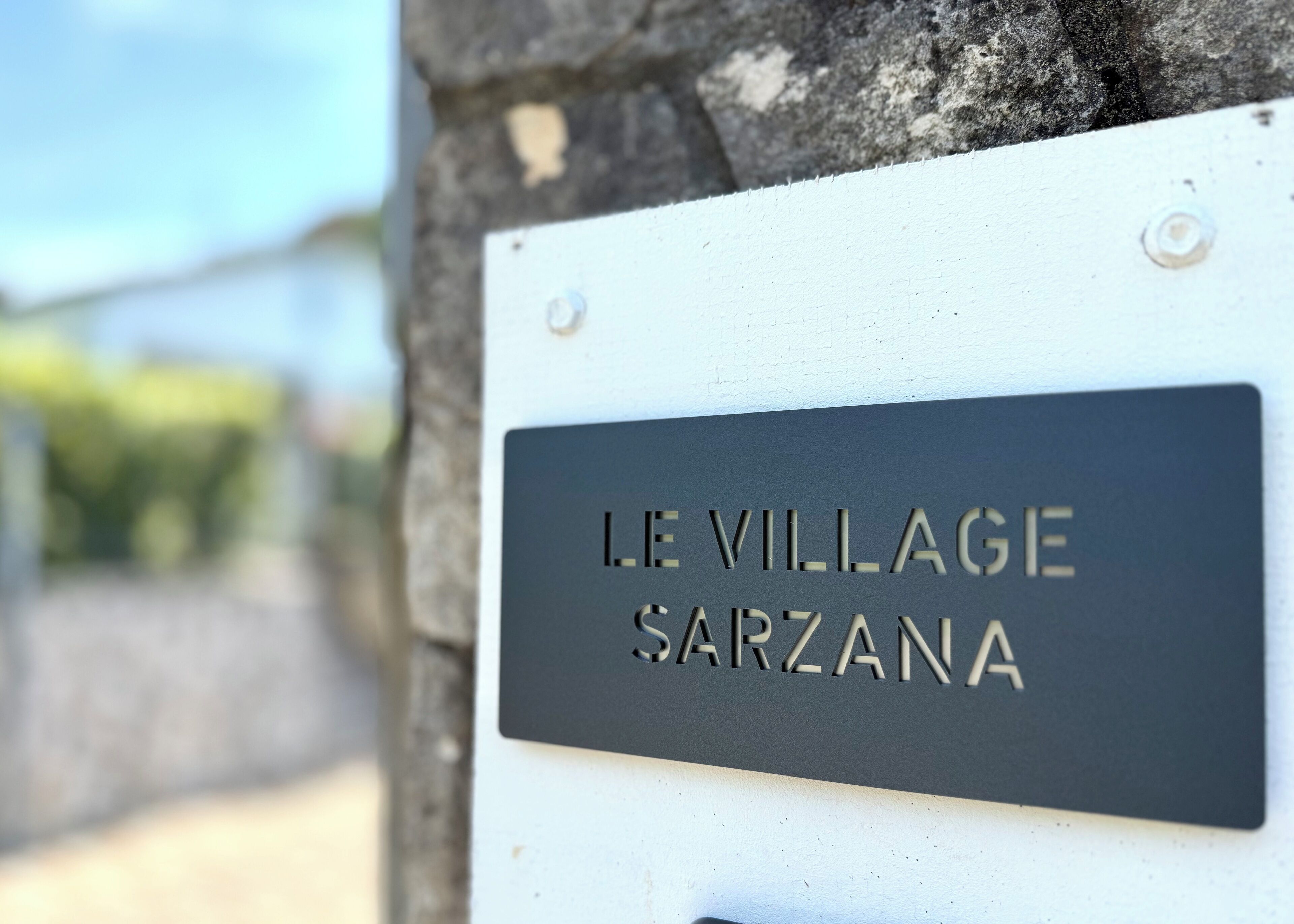 A sign with the text "LE VILLAGE SARZANA" mounted on a stone wall.