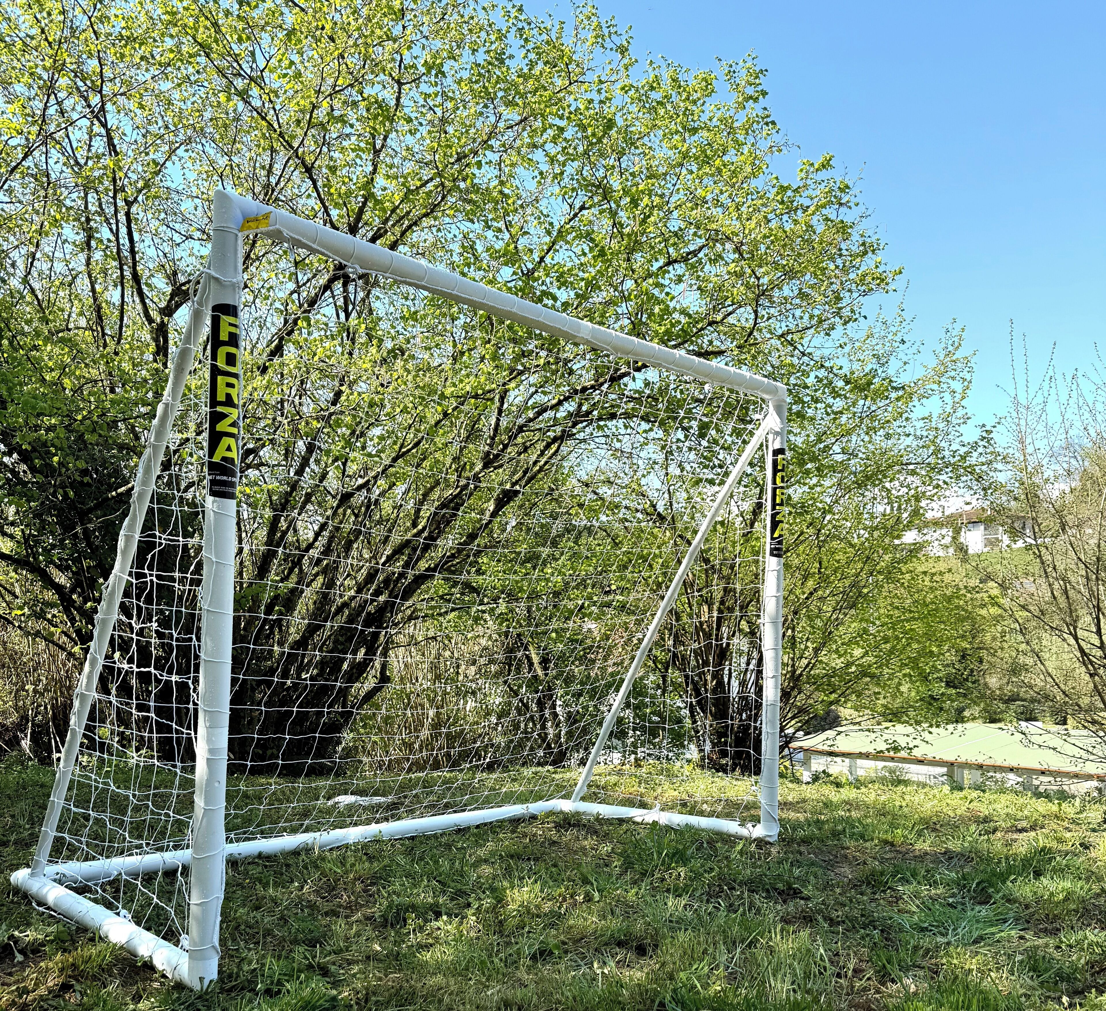 Un but de football portant la marque « FORZA » sur les poteaux, installé dans une zone herbeuse avec des arbres en arrière-plan.