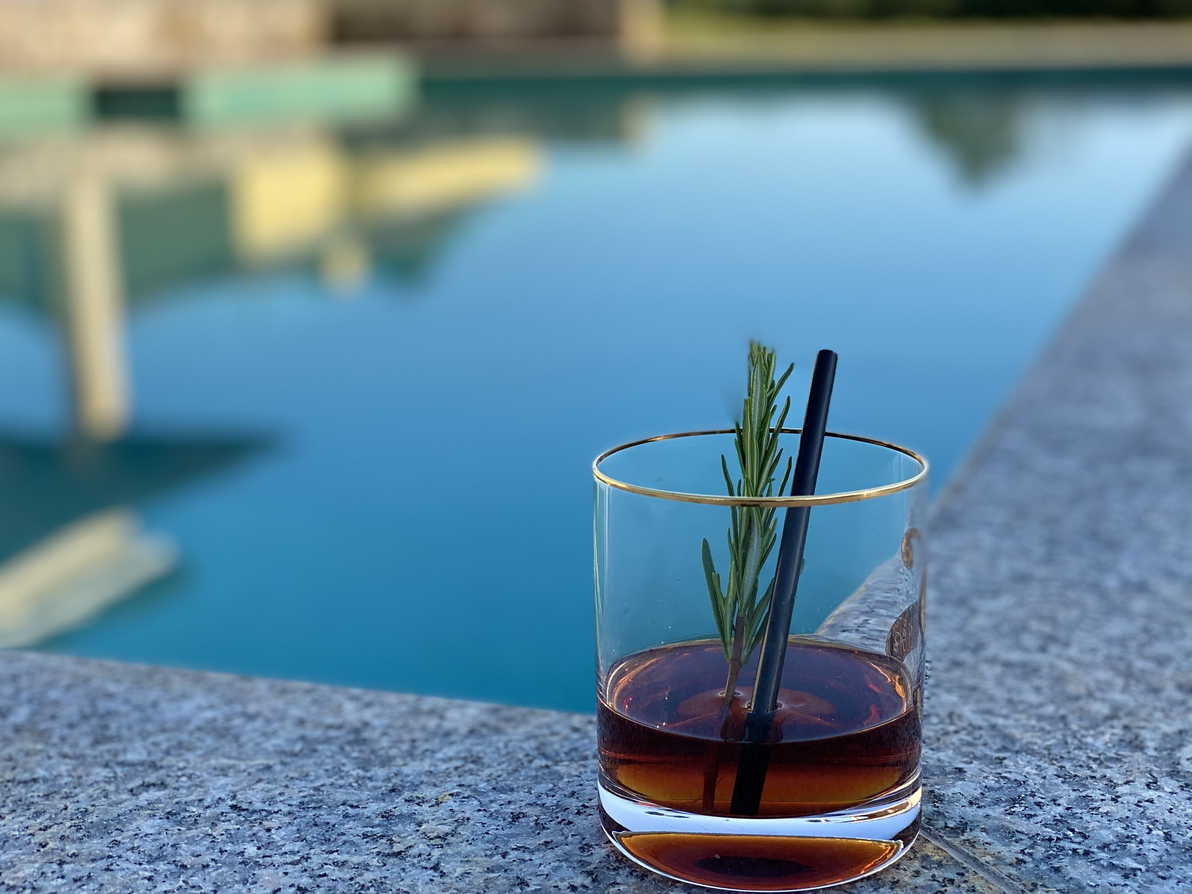 Et glas med gyldent væske, rosmarin og et sugerør, placeret på en stenoverflade ved en pool.