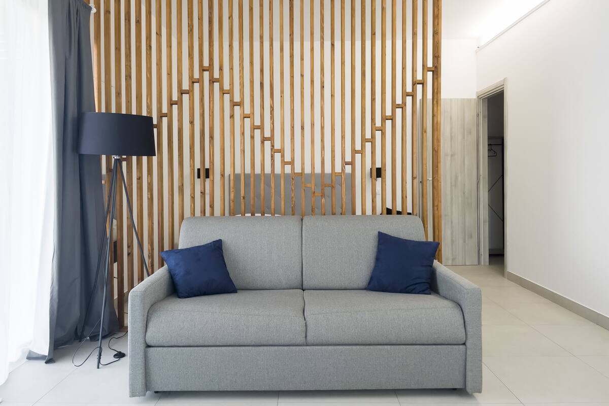 Een moderne woonkamer met een grijze bank, twee blauwe kussens, een vloerlamp en een houten lattenwand.