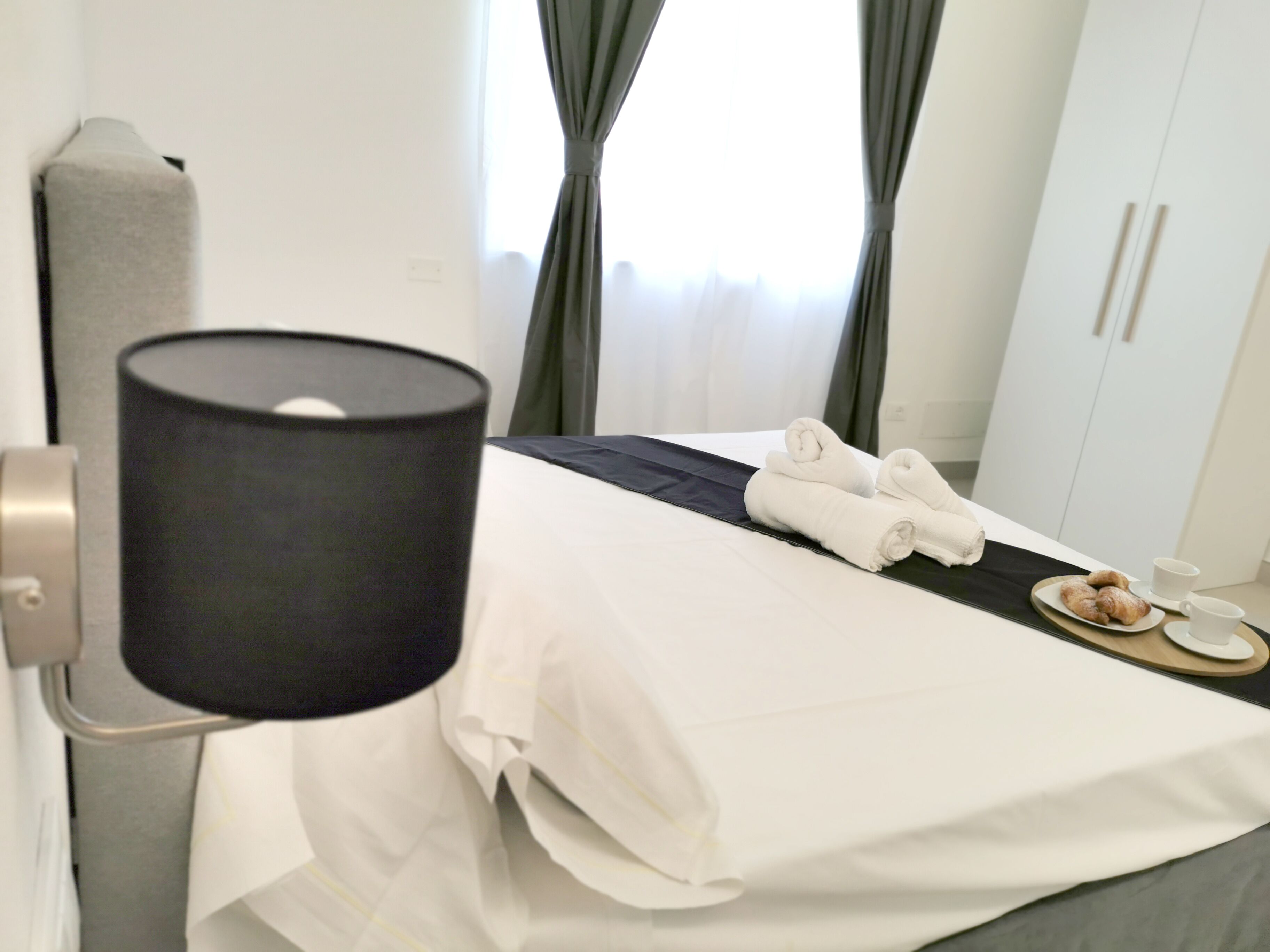 Une chambre d’hôtel avec un lit, une lampe de chevet, une paire de serviettes et un plateau de petit-déjeuner avec des croissants et une tasse.