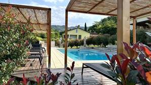 Zona de piscina exterior con una pérgola, sillones de descanso y vistas a una casa y colinas al fondo.