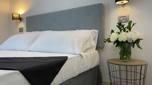 Una cama bien hecha con sábanas blancas y una manta negra, un cabecero tapizado en gris y un florero con flores blancas sobre una mesita redonda.