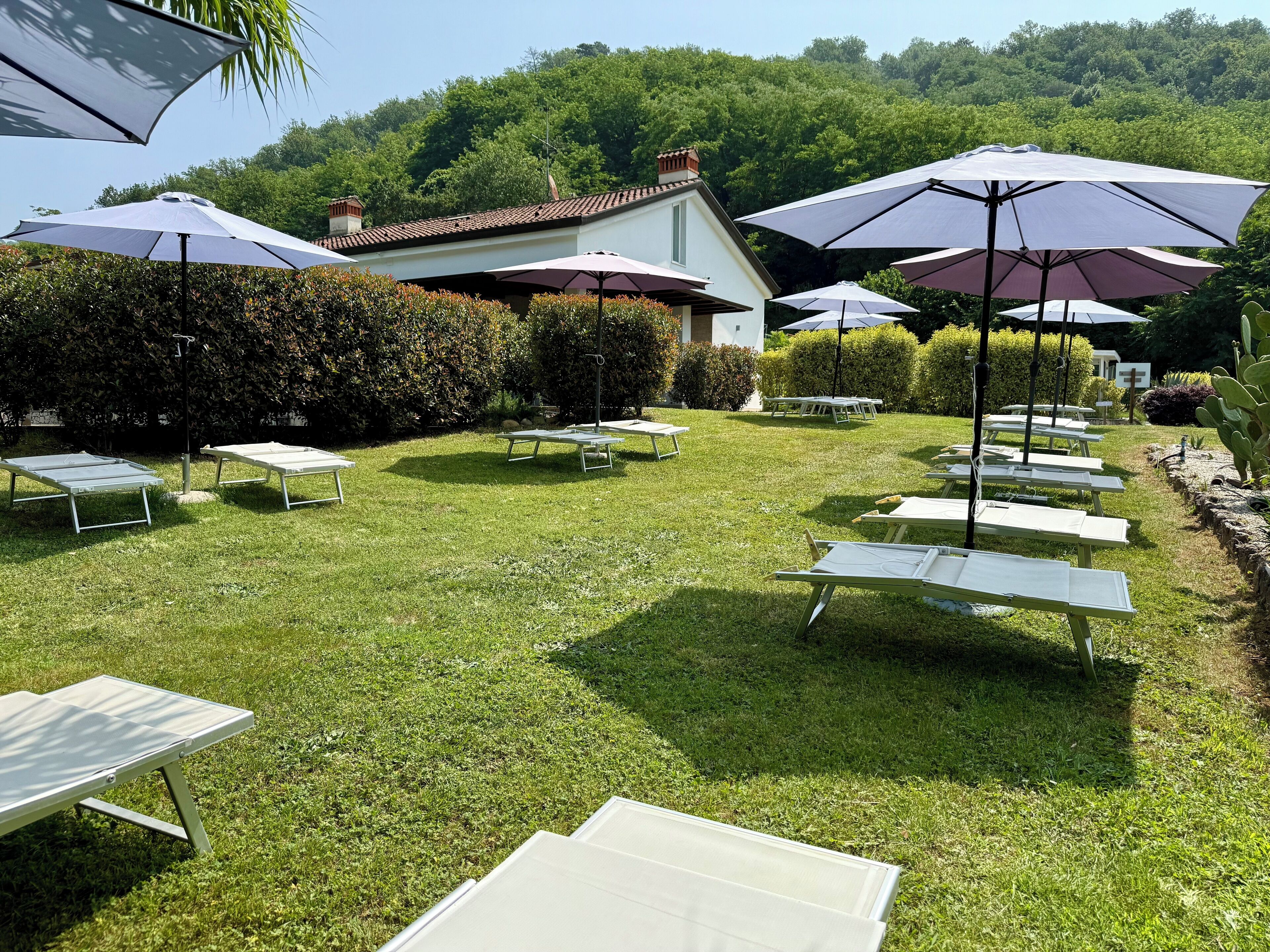 An outdoor seating area with tables and umbrellas, surrounded by greenery and a building in the background.