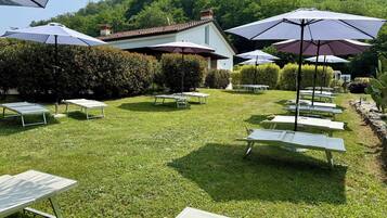 An outdoor seating area with tables and umbrellas, surrounded by greenery and a building in the background.