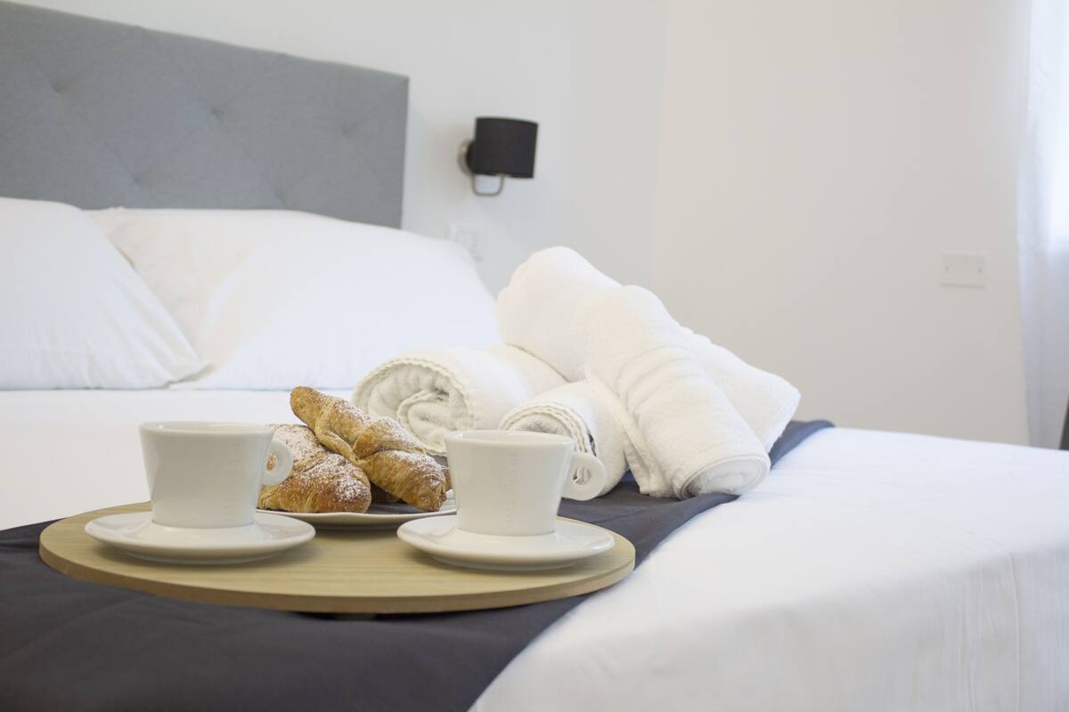 Een net opgemaakt bed met witte lakens, een dienblad met croissants en koffiekopjes, en een wandlamp.